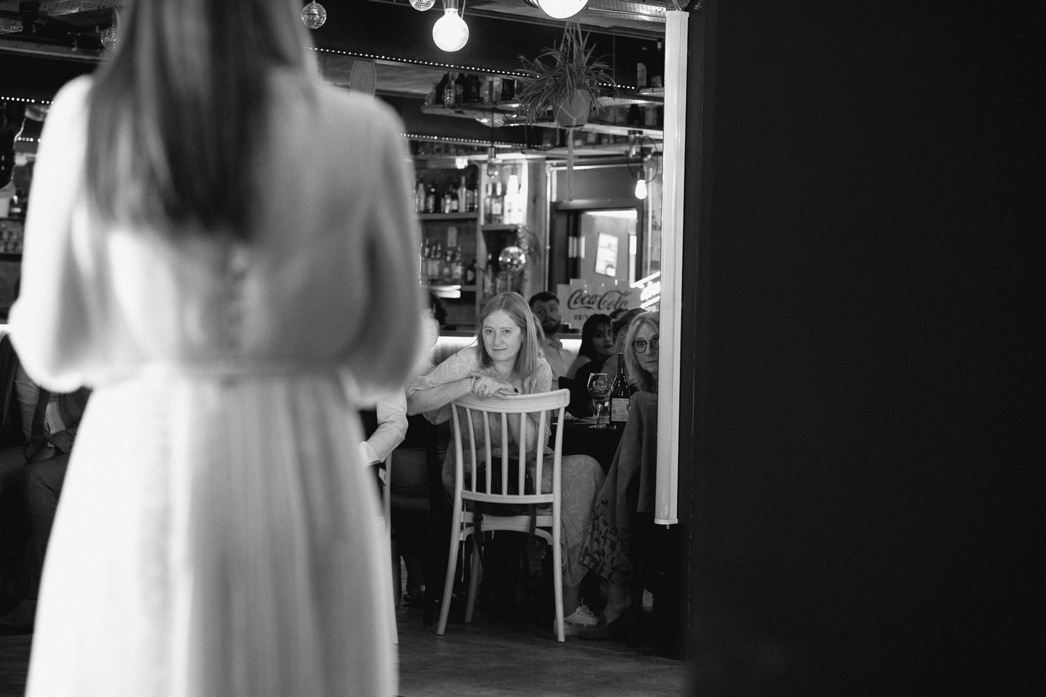 Black and white photo of a woman in a white dress standing in a doorway of a cozy restaurant.