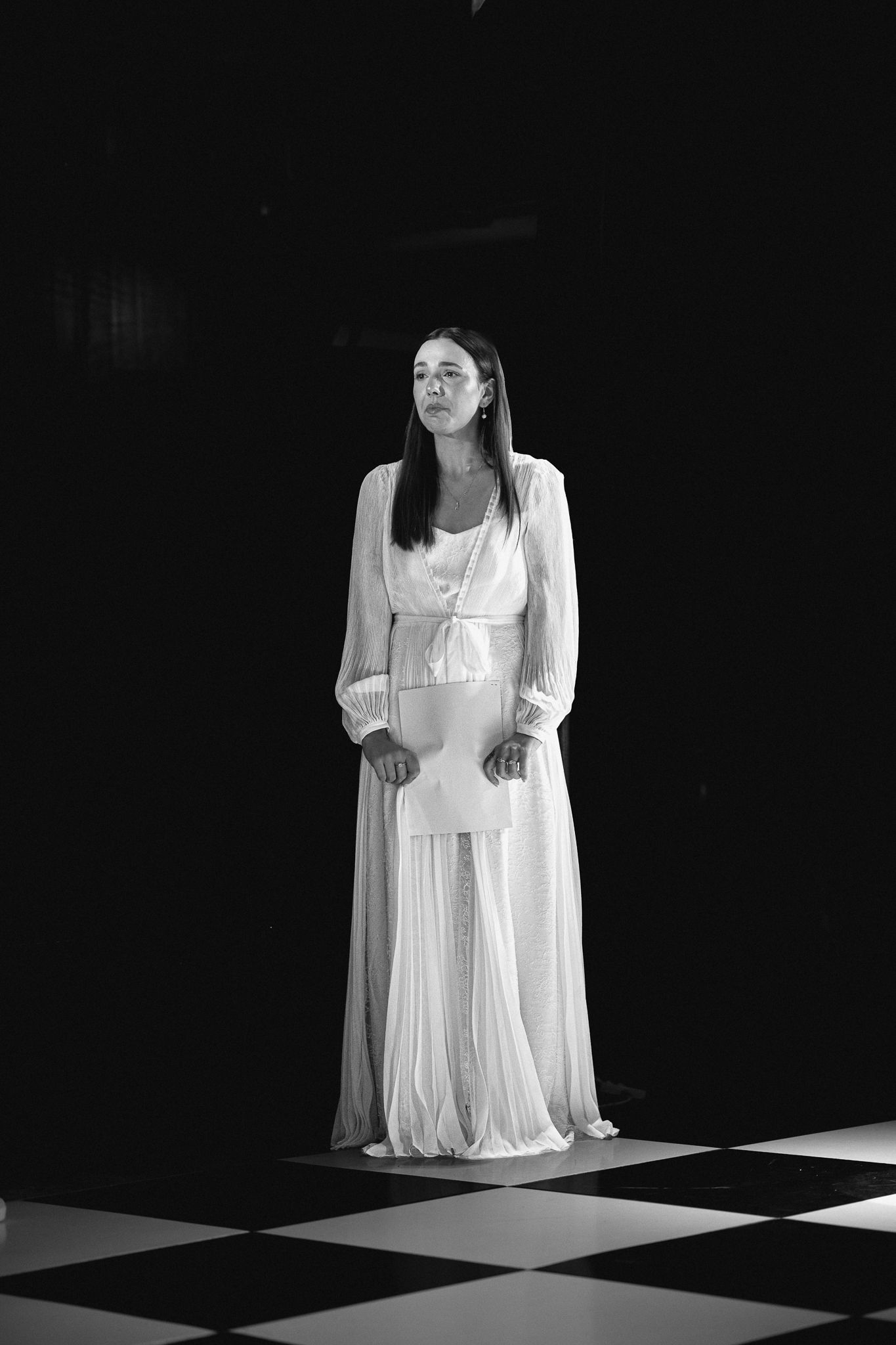 Woman in white flowing gown standing on checkered floor in dramatic black and white portrait photography.