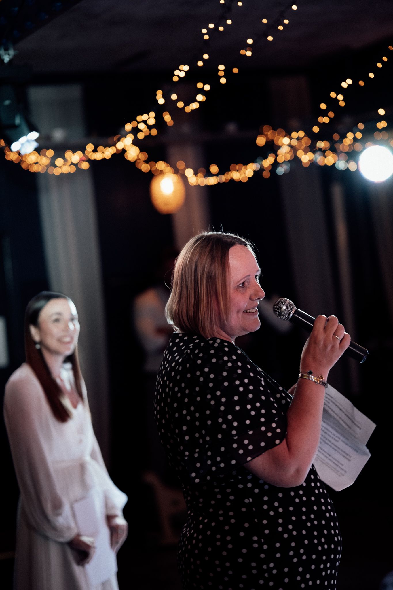 Woman in polka dot dress speaking into microphone at evening event with bokeh string lights backdrop.