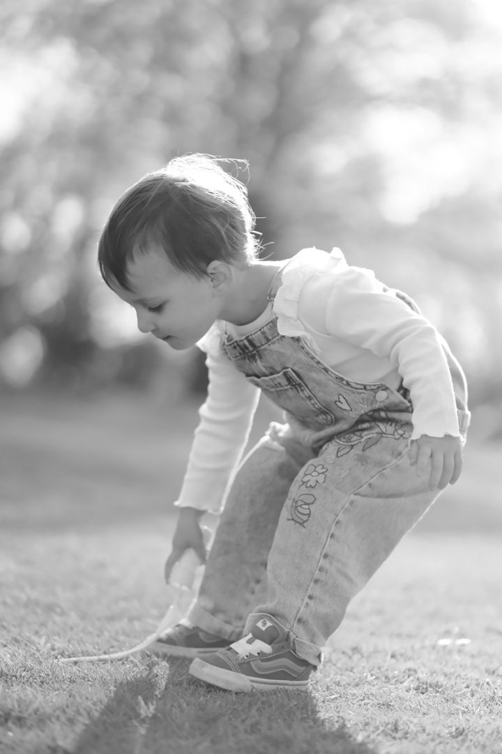 Small child in denim and white crouches down to explore something on the grassy ground in black and white.