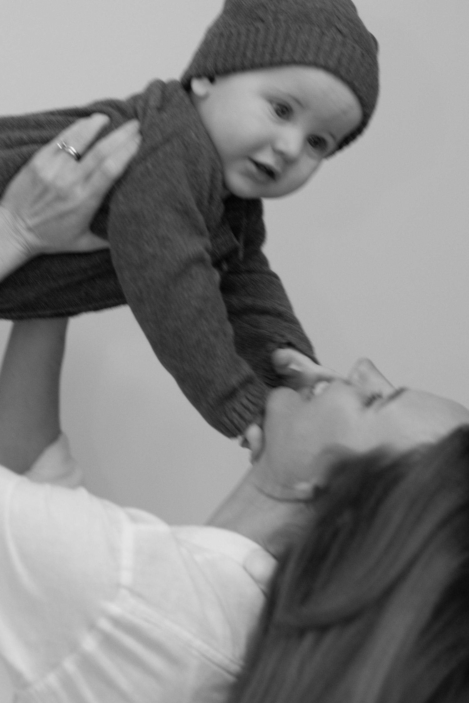 Black and white photo of a mother lifting her smiling baby in a knit hat above her face in a joyful moment.