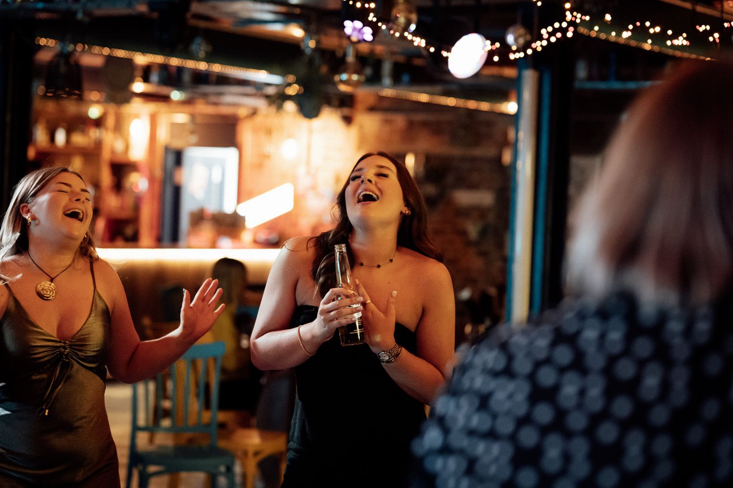 Two women singing karaoke passionately at a lively bar, one holding a microphone under warm string lights.