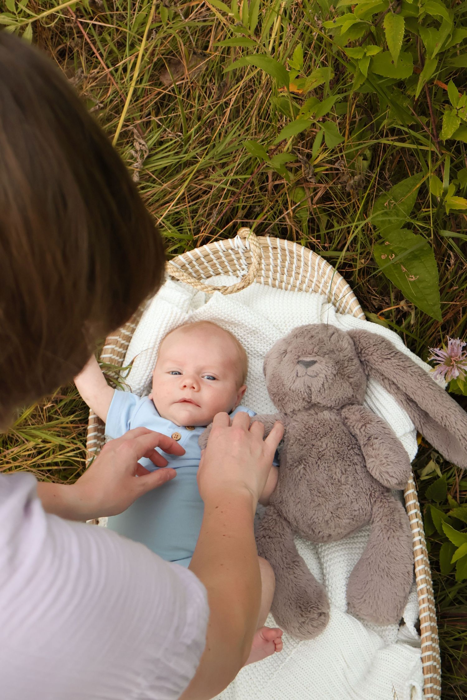 A baby lies on a white basket with a plush bunny toy in an outdoor garden setting.
