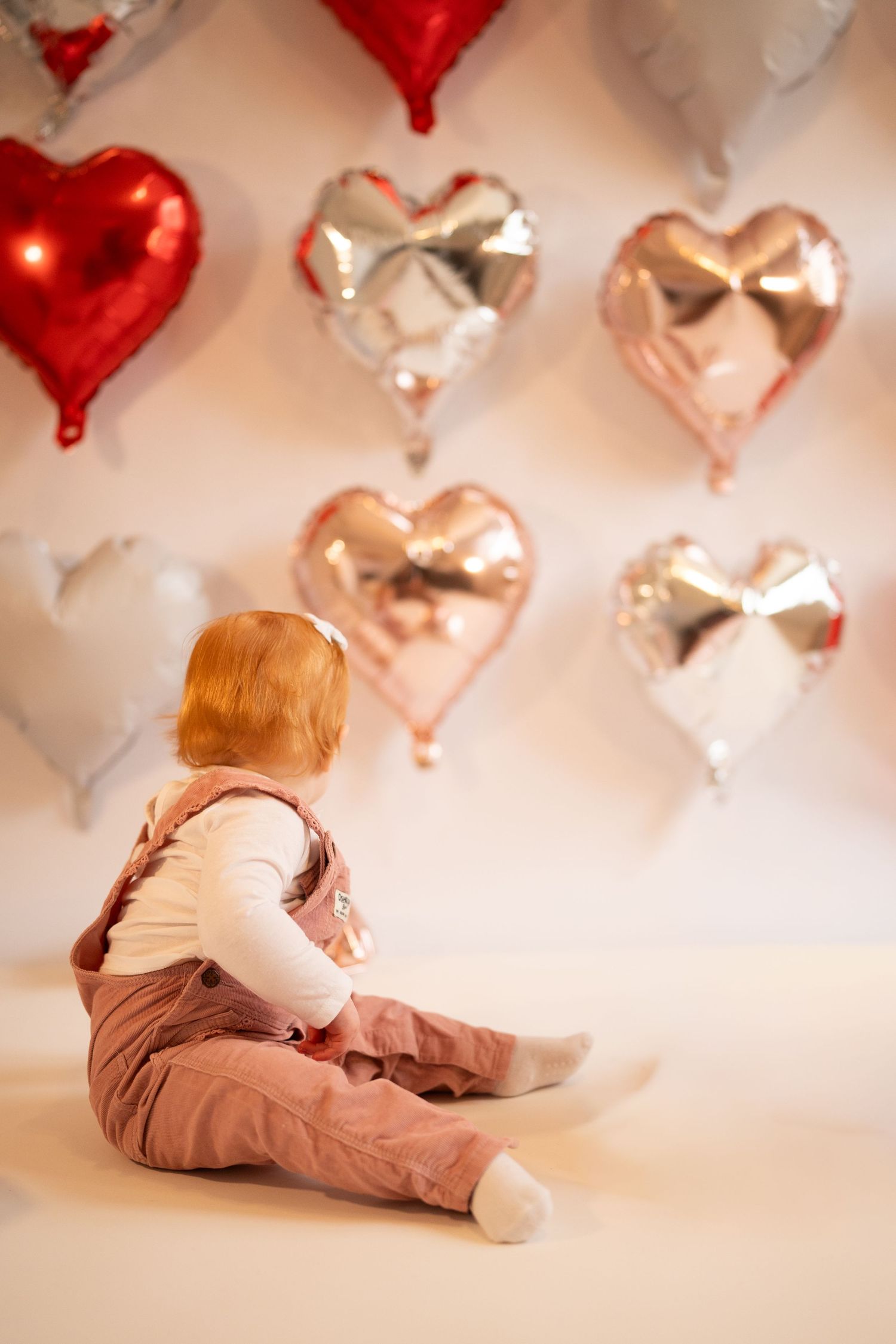 A child lies on a white floor with red heart-shaped balloons floating above in a dreamy Valentine's Day setting.