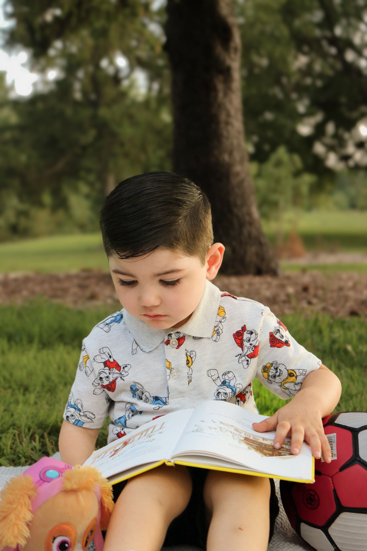 Child in a patterned shirt reading a book while sitting on grass.