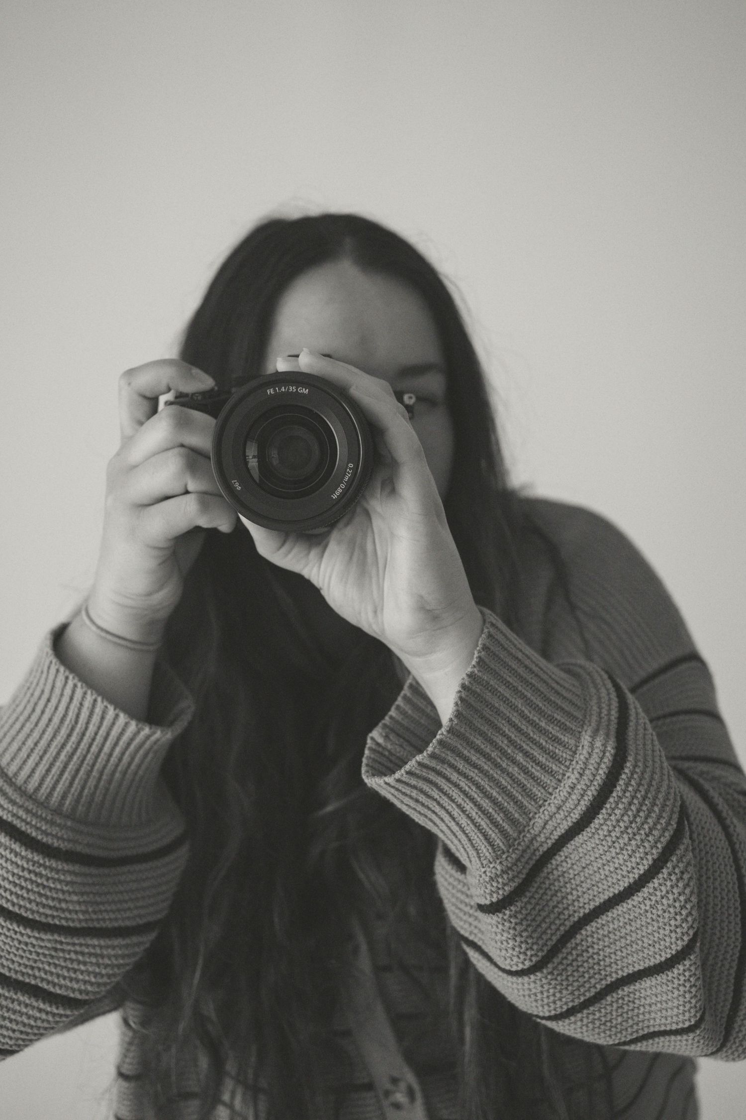 Black and white photo of a woman with long hair holding a DSLR camera up to her face while wearing a striped sweater.