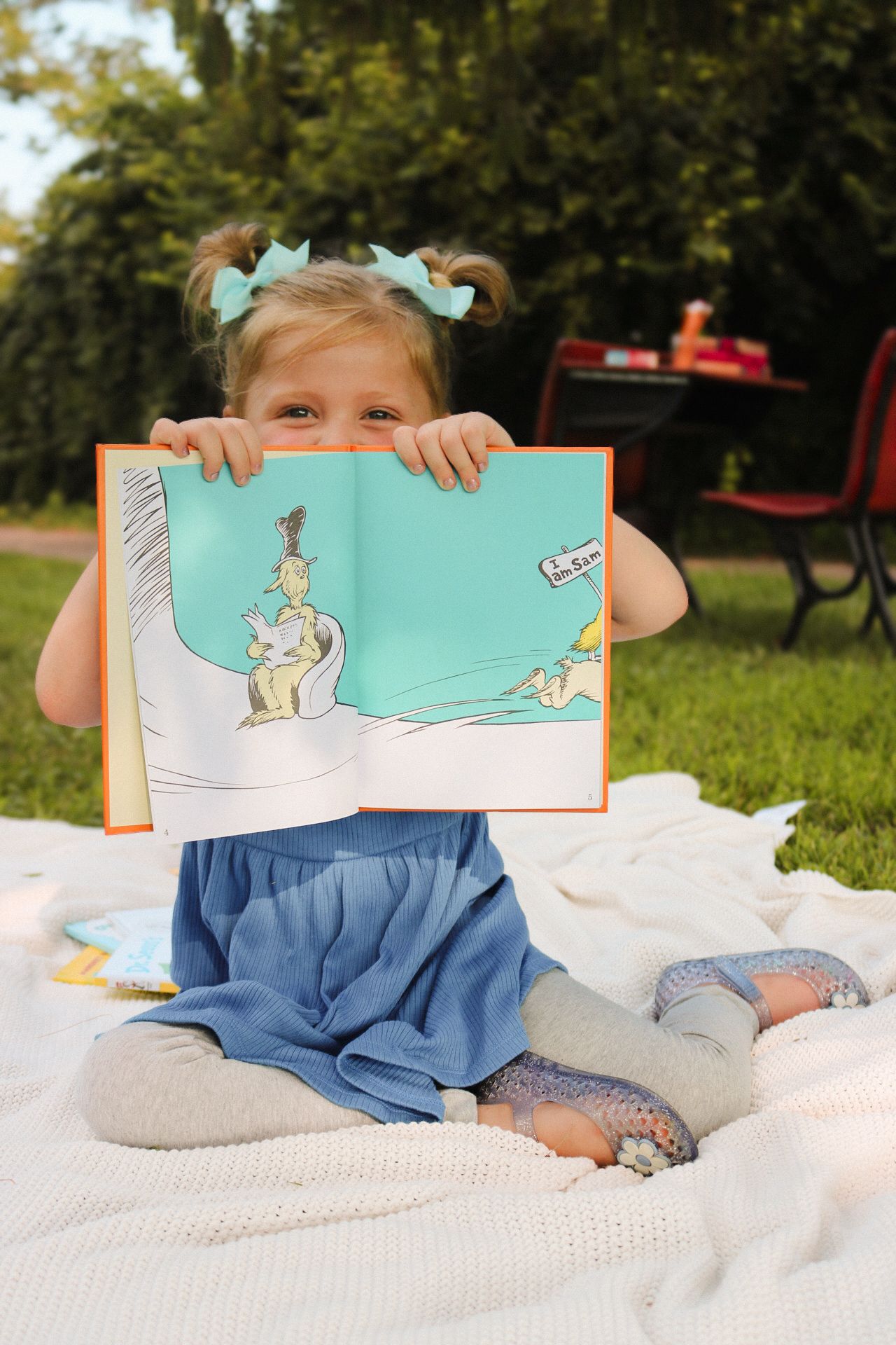 Small child reading a picture book while sitting on a white blanket outdoors.