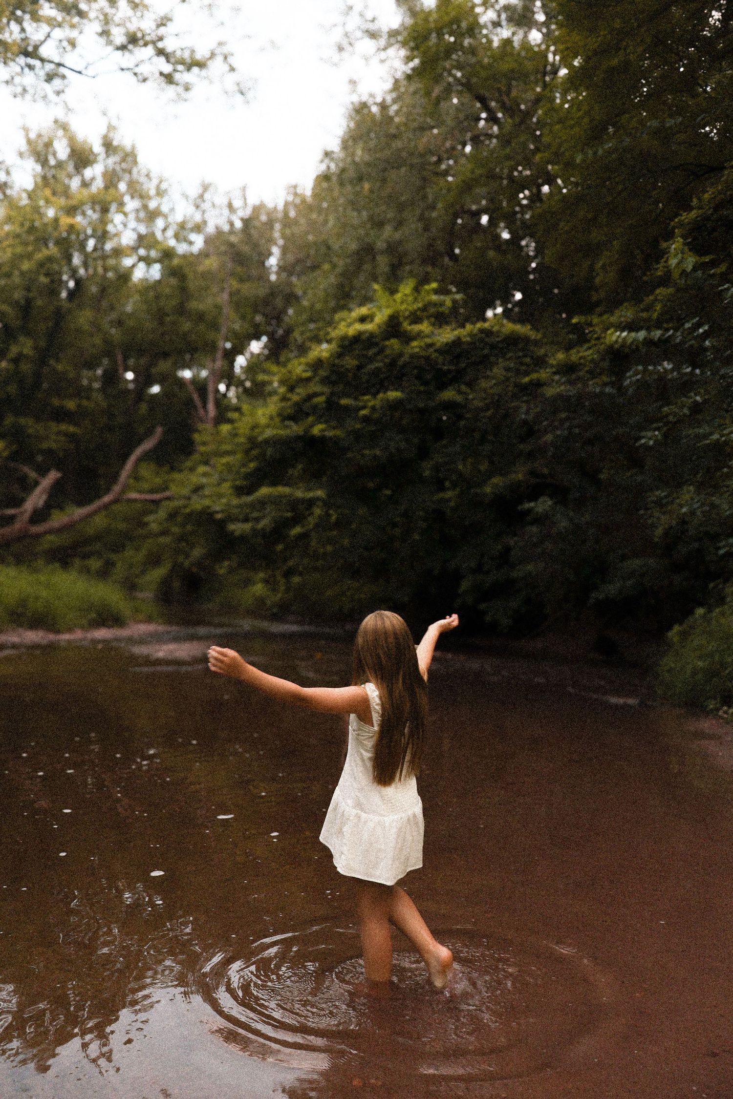Person in white dress splashing and playing in brown river water at dusk.