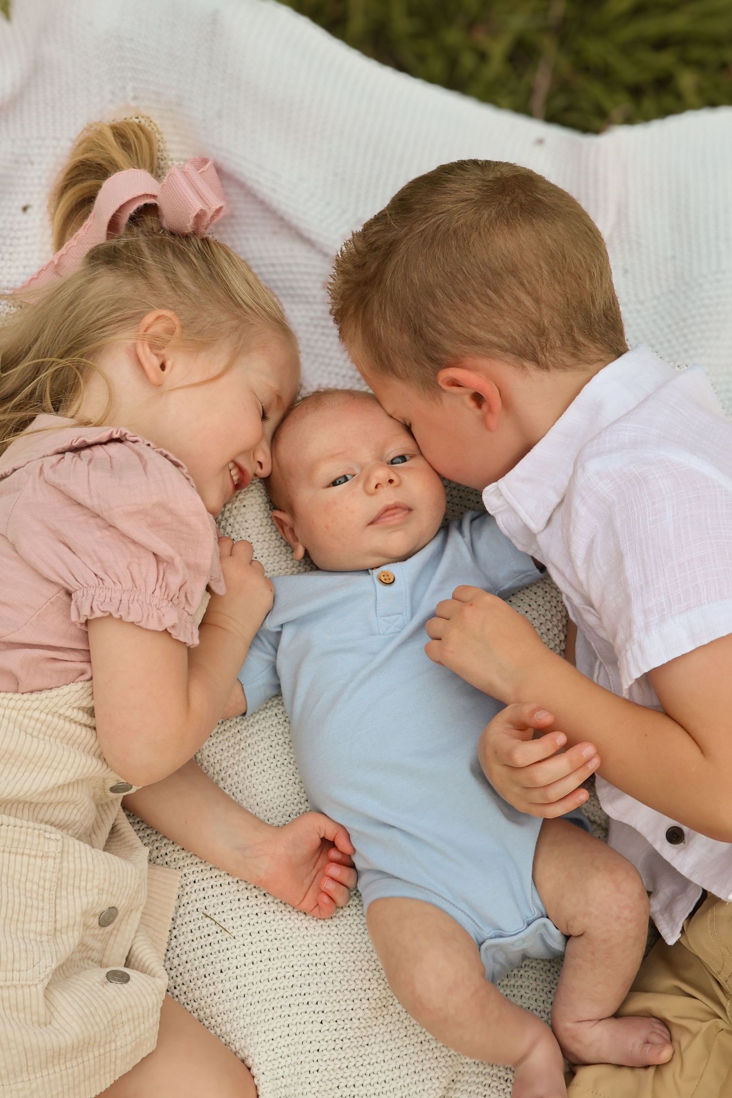 Three young siblings sharing tender moment on white blanket outdoors.