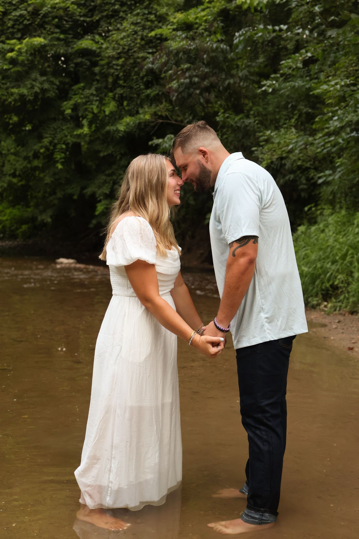 A couple stands in a shallow creek together wearing white and dark clothing in a romantic moment.