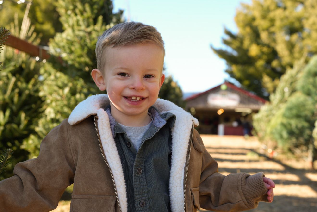 A cheerful toddler in a brown jacket with white fleece collar smiles outdoors on a sunny autumn day.