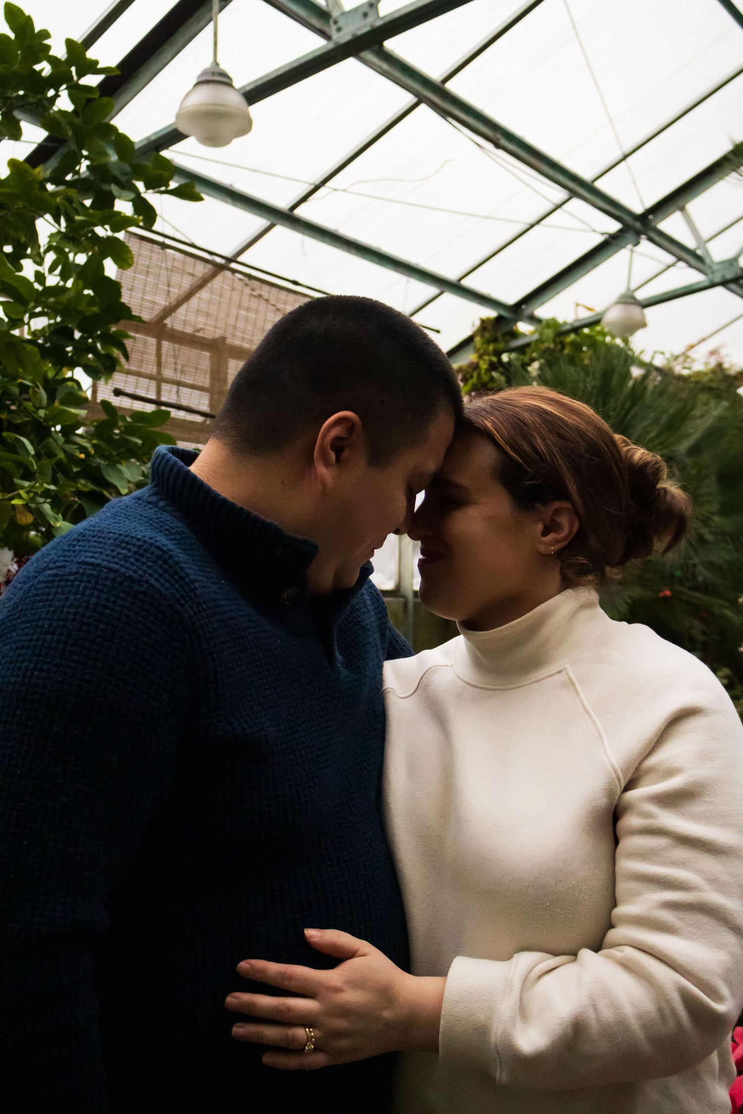 Two people in a greenhouse share an intimate moment wearing blue and white sweaters.