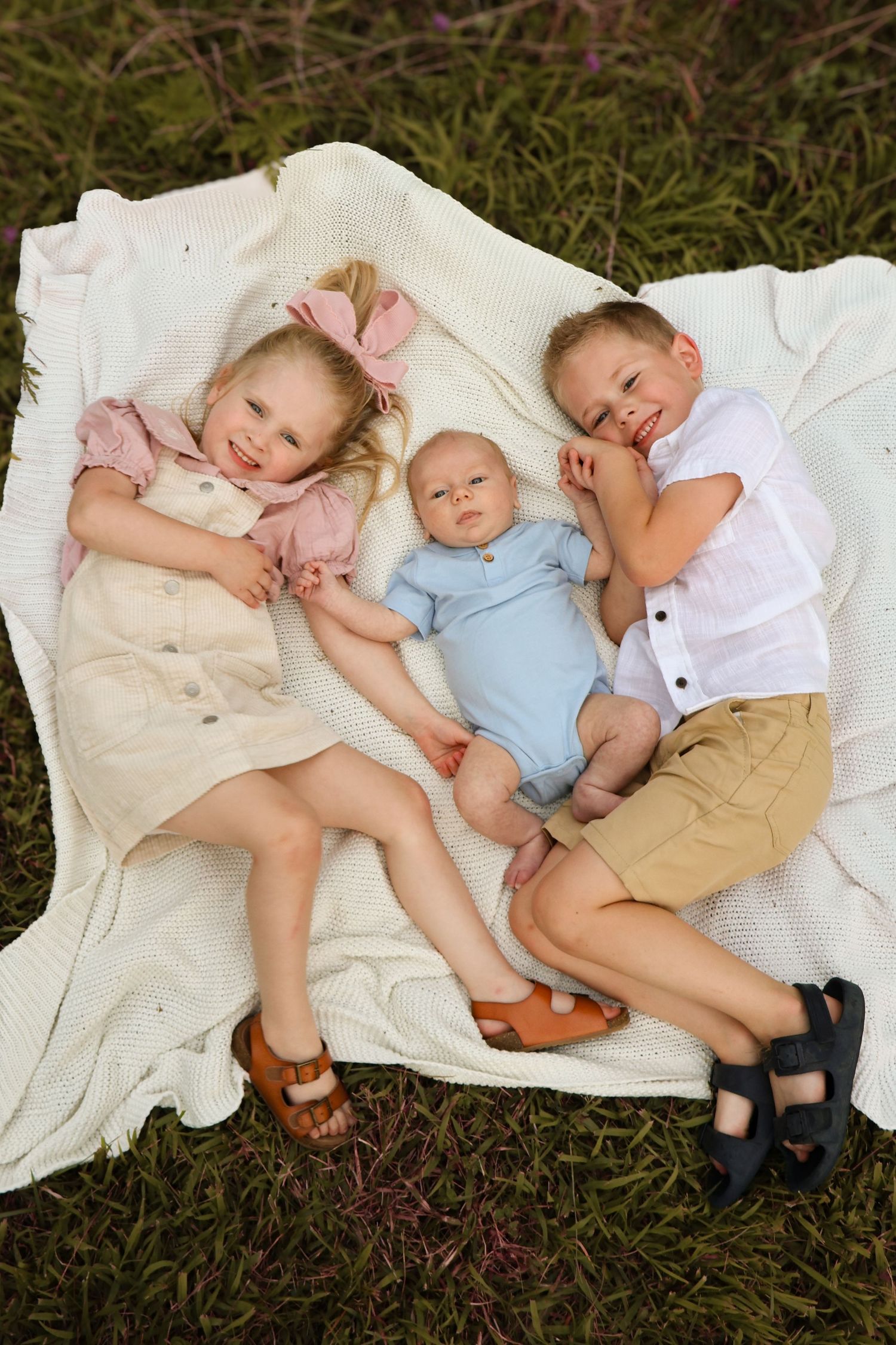 Children resting together on a white blanket in the grass during an outdoor photo session.