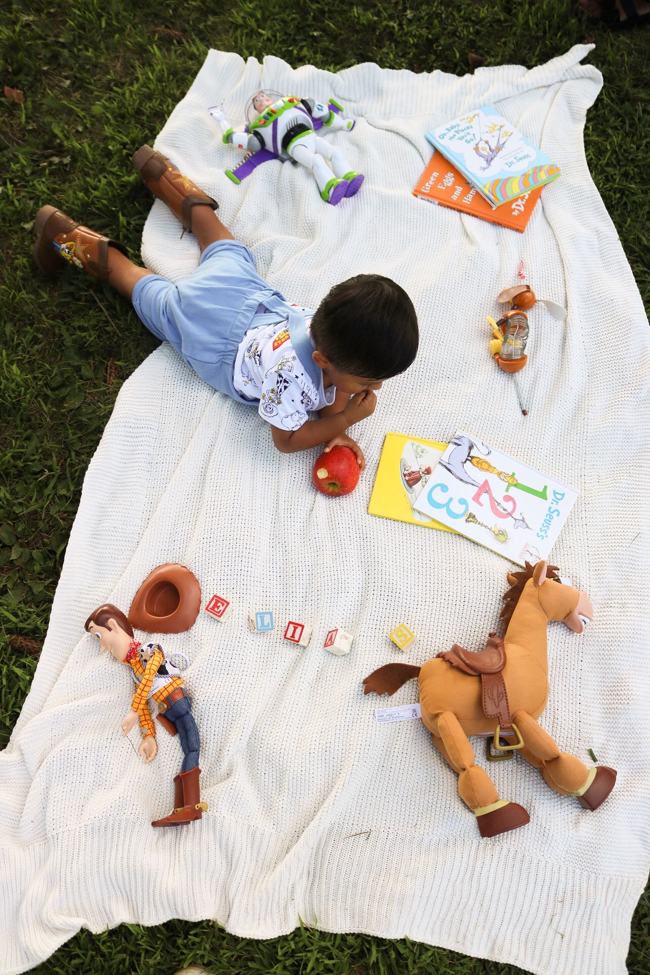 Children lying on white blanket reading books and coloring during outdoor activity.