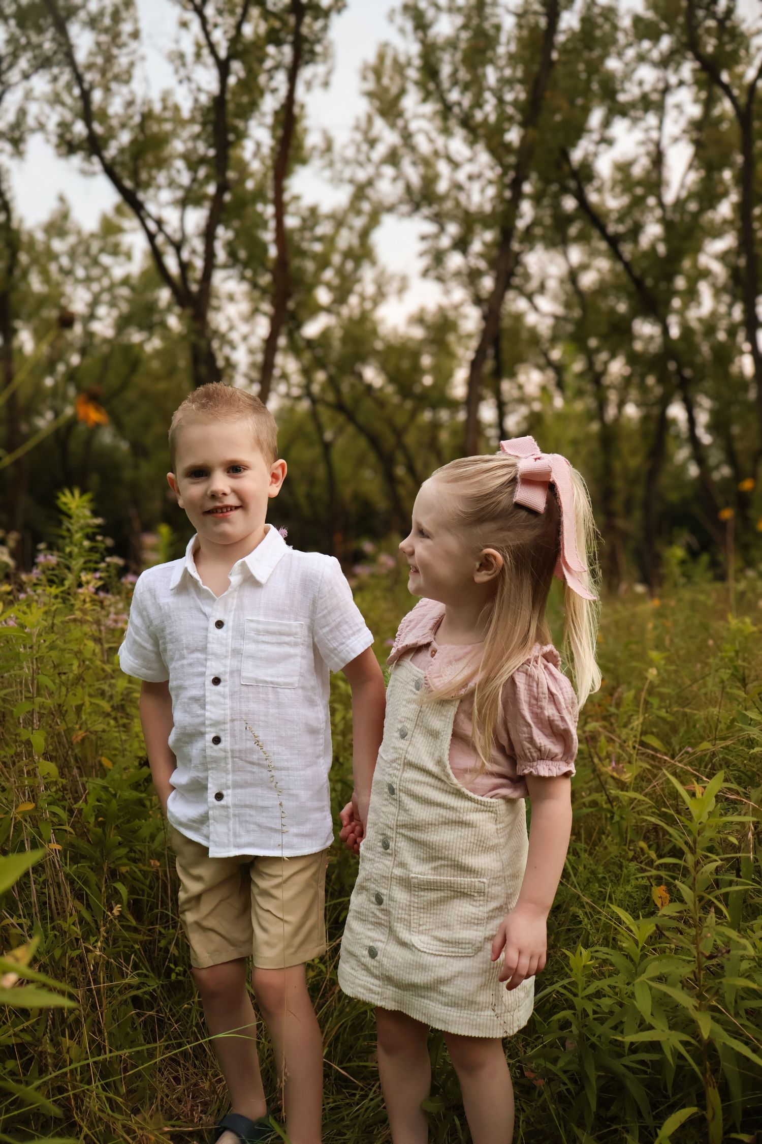 Two children stand together in tall grass with trees in the background.