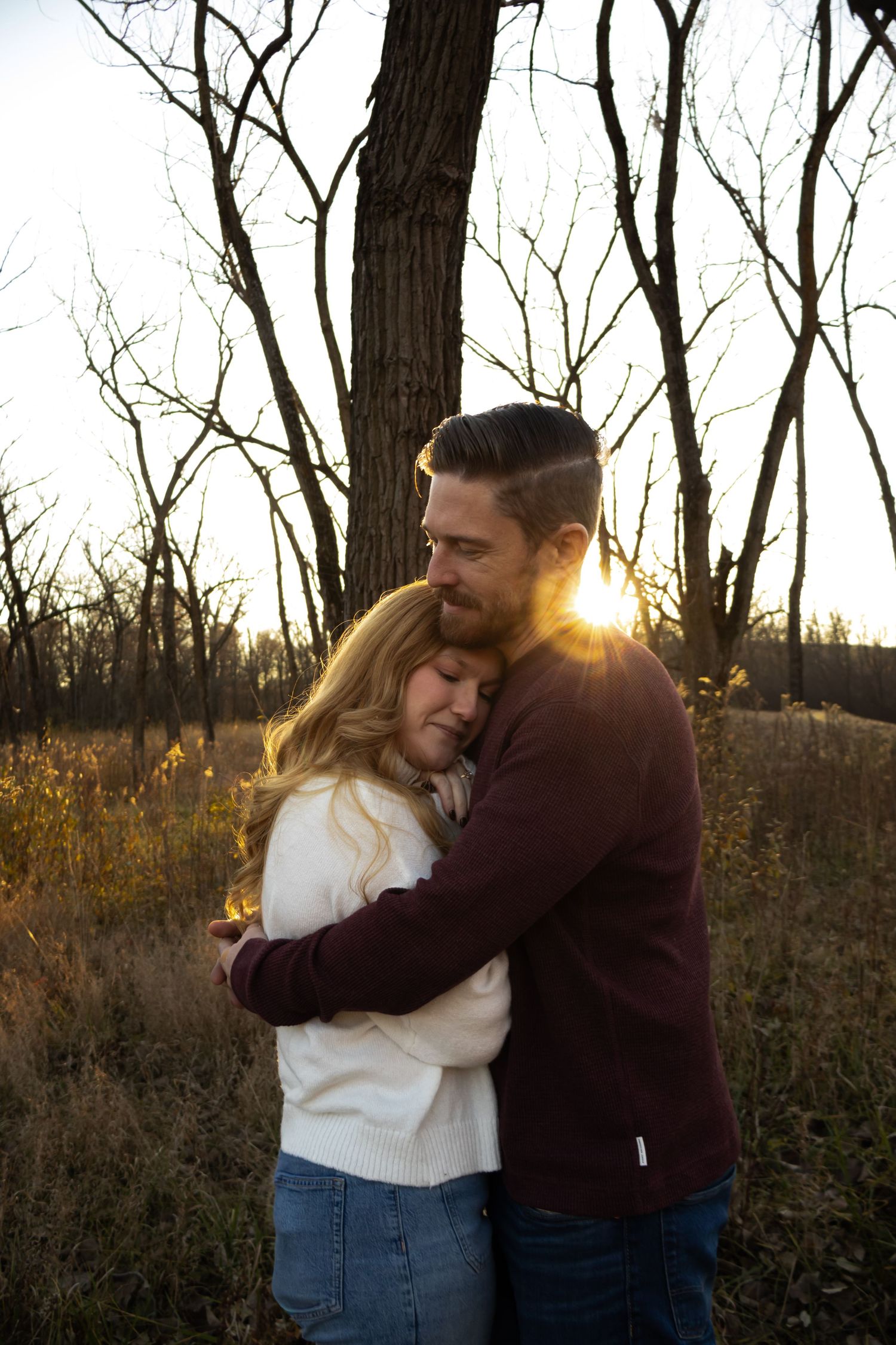A couple embraces in a romantic outdoor setting during golden hour with bare trees and tall grass in the background.