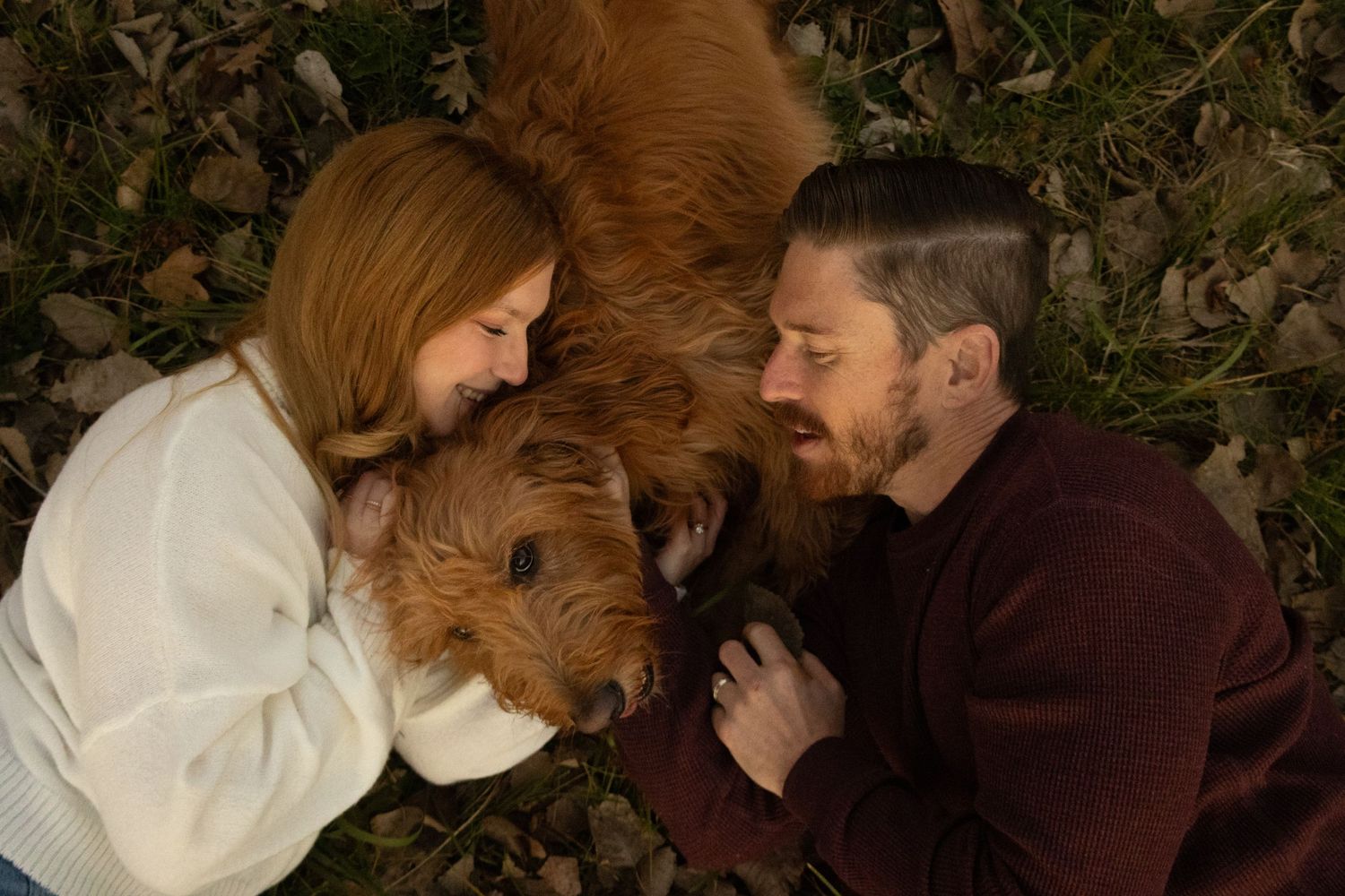 A couple lies on autumn leaves with their golden retriever dog between them.