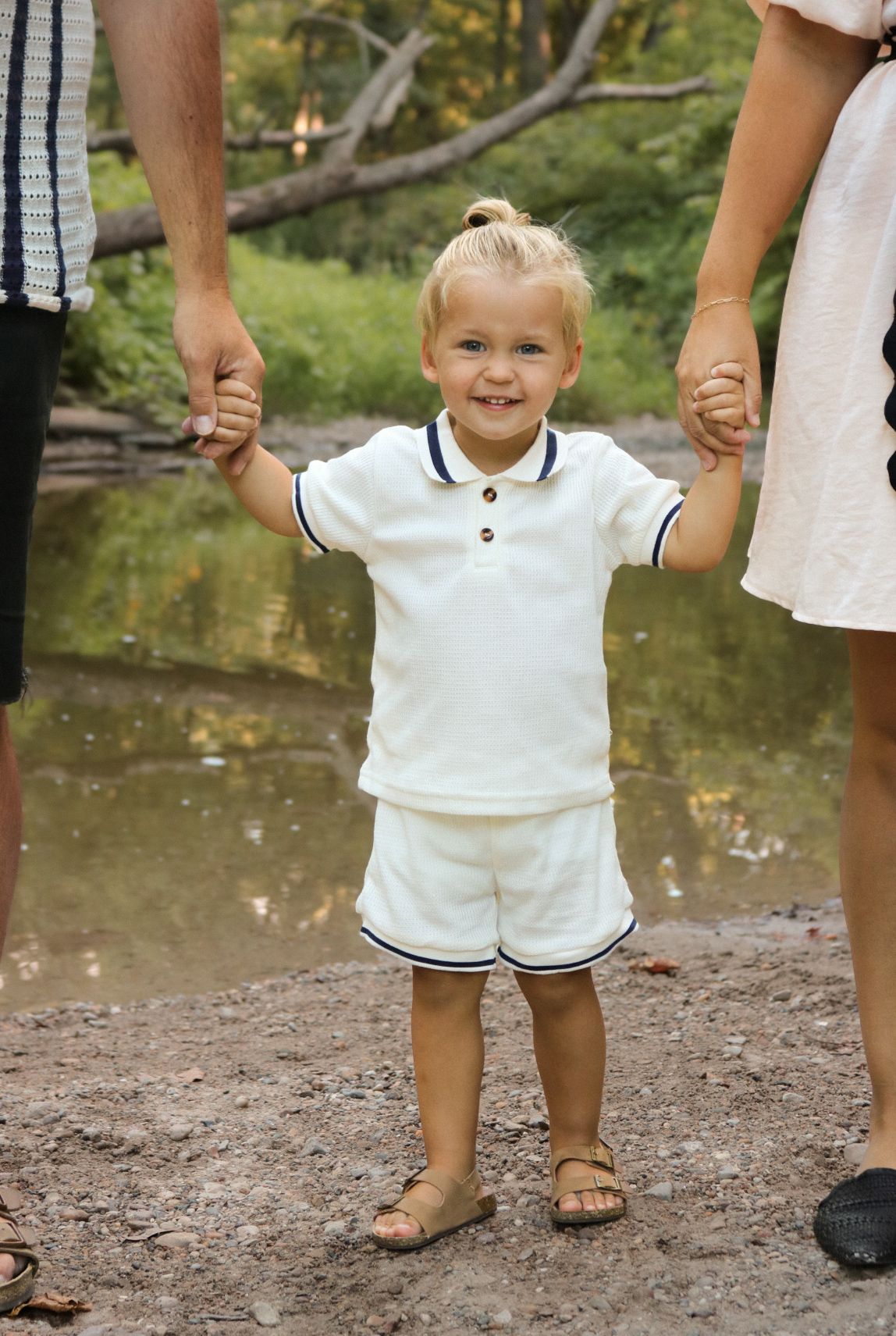 Small child in white outfit holding hands with adults by a creek.