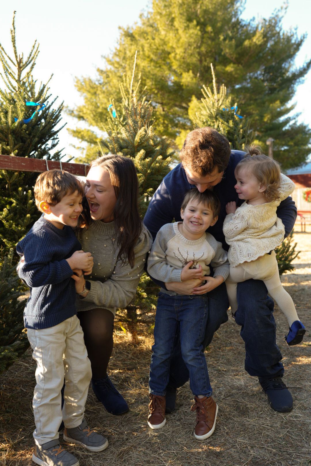 A family shares joyful moments together while standing among evergreen trees at a Christmas tree farm.
