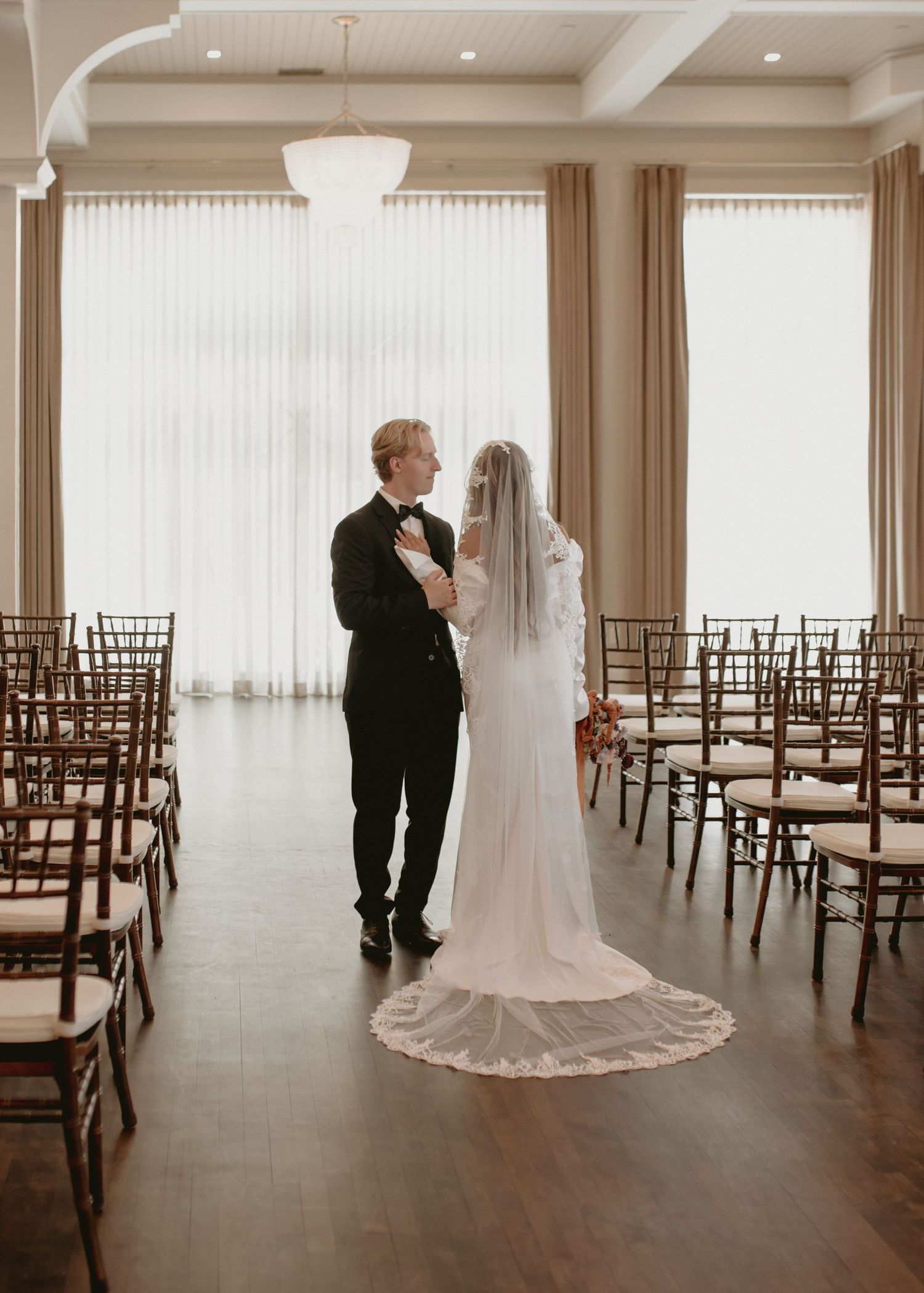 Newlyweds share an intimate moment in an elegant reception hall with wooden chairs and natural light.