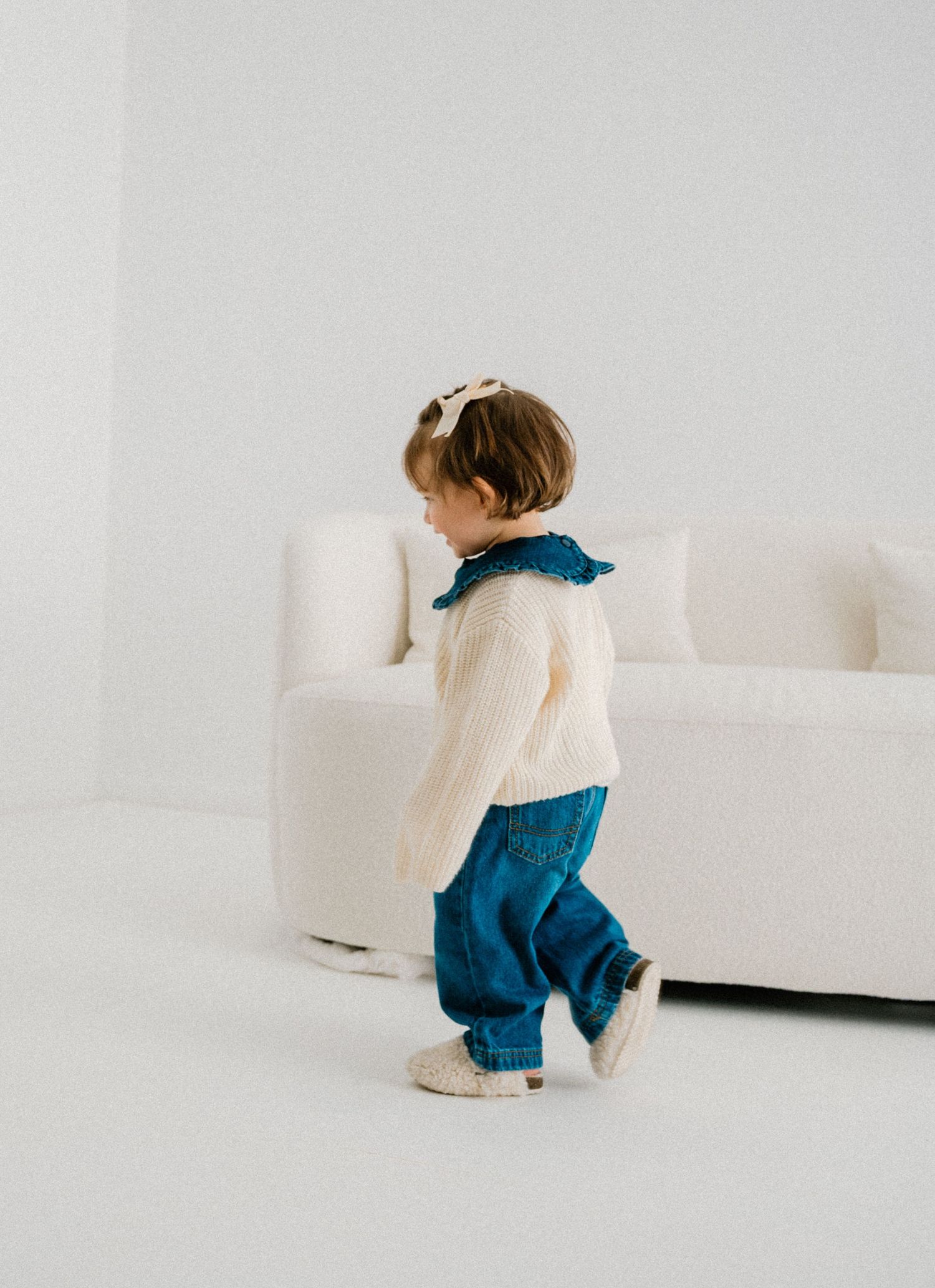 Baby wearing cream sweater and blue pants crawling on white couch. Appleton, Green Bay & Kaukauna