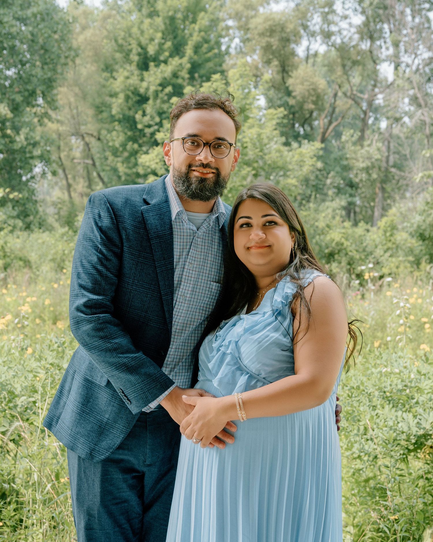 A couple embraces while standing in a meadow with trees, wearing blue formal attire on a sunny day. Appleton, Green Bay & Kaukauna