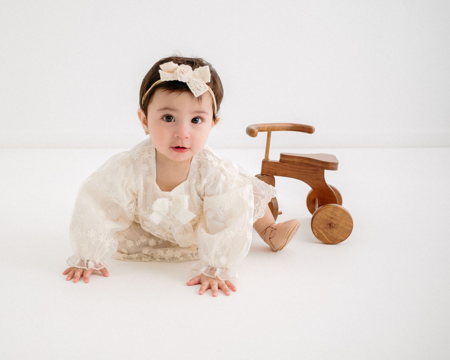 Baby in white lace dress crawling near wooden toy on white background. Appleton, Green Bay & Kaukauna