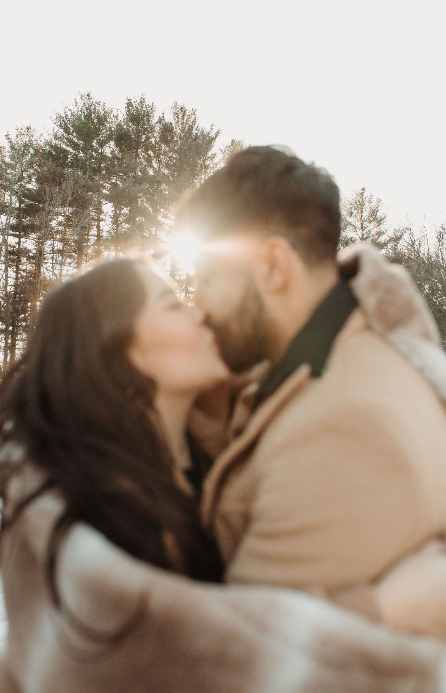 Couple embraces tenderly during a golden hour photoshoot with sunlight streaming through trees in the background.