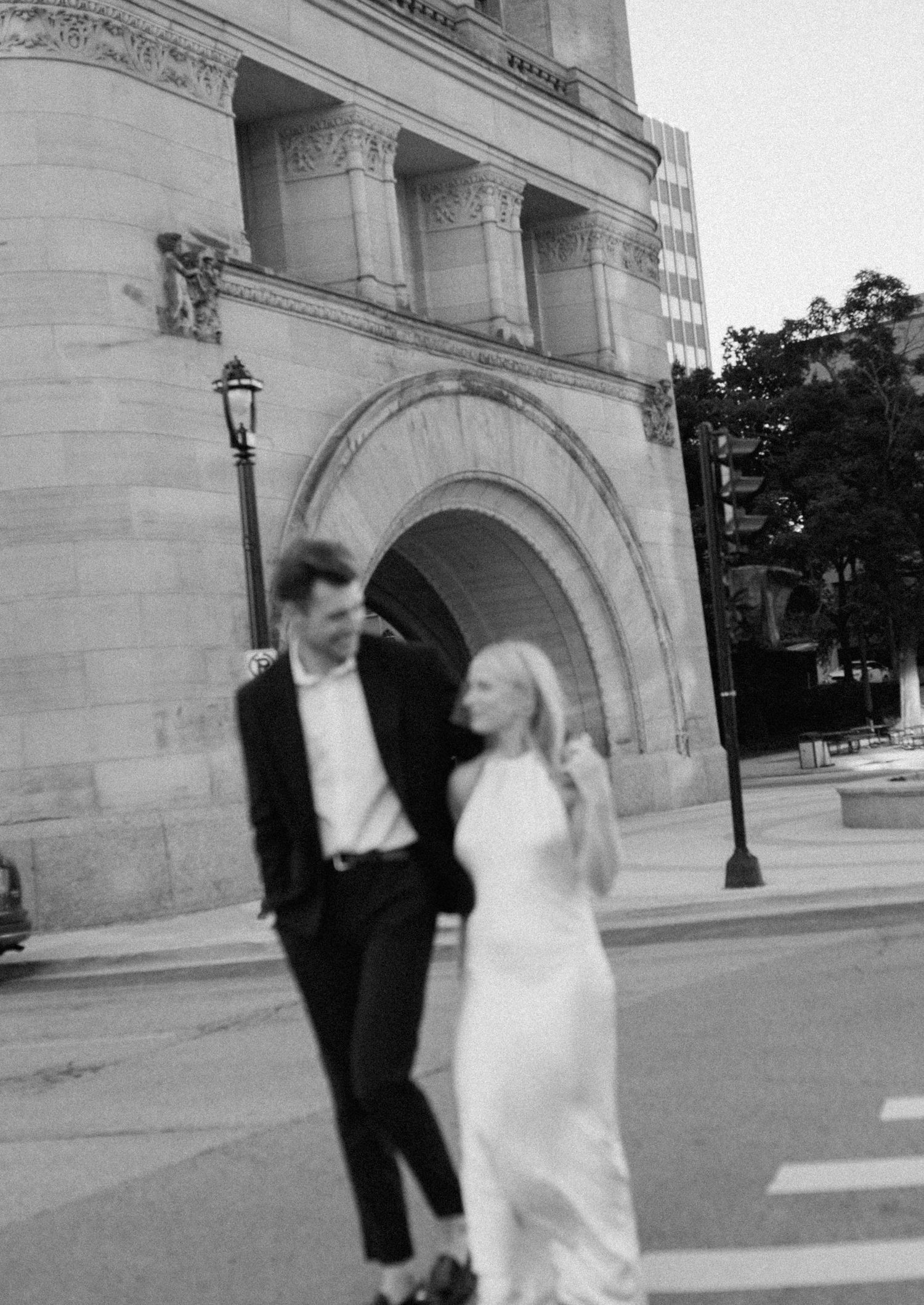 Black and white photo of couple sharing a moment outside stone building with arched entrance.