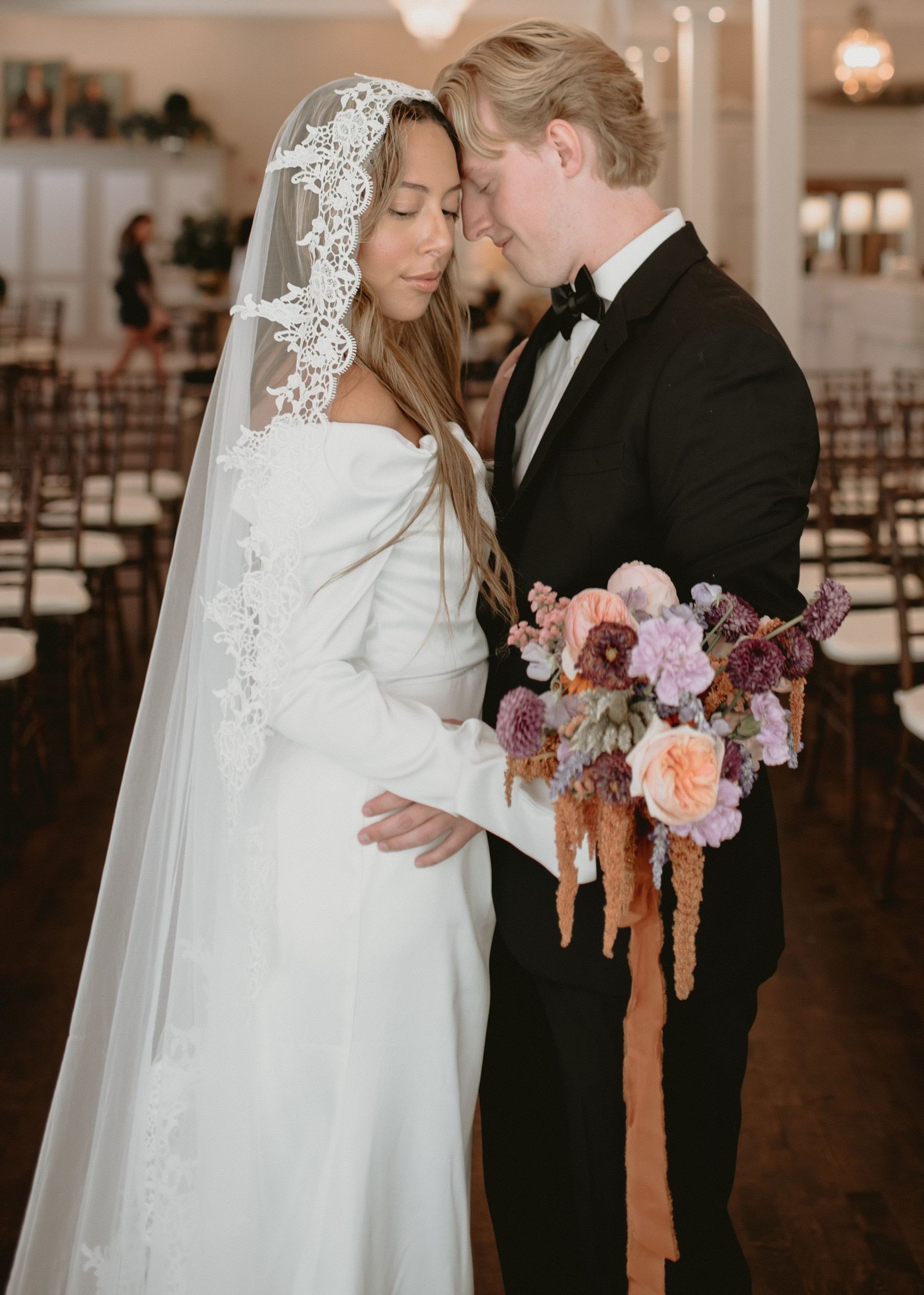 Wedding couple sharing intimate moment with bridal bouquet in elegant indoor venue.