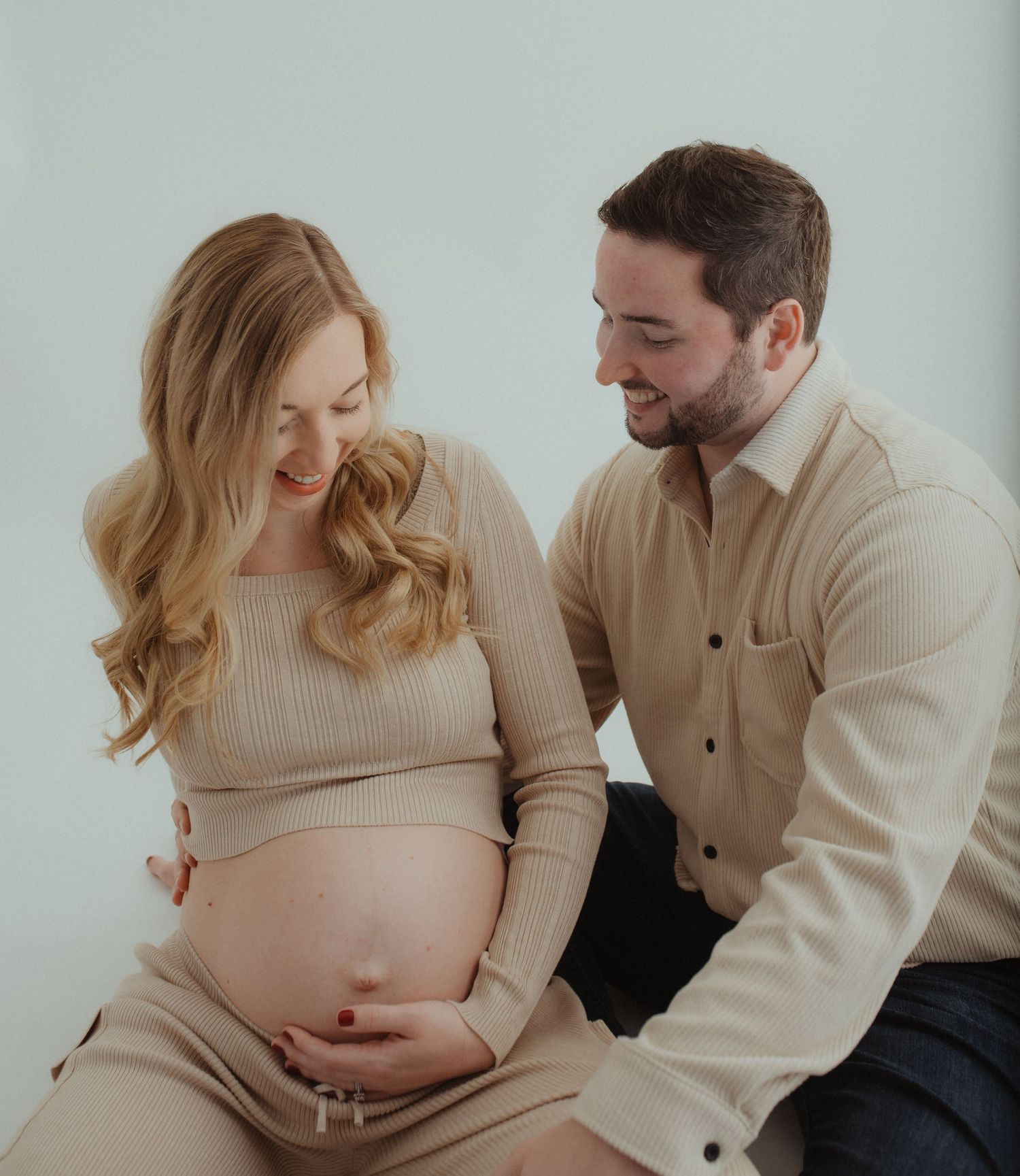 Intimate maternity photoshoot with couple in neutral sweaters against light background.