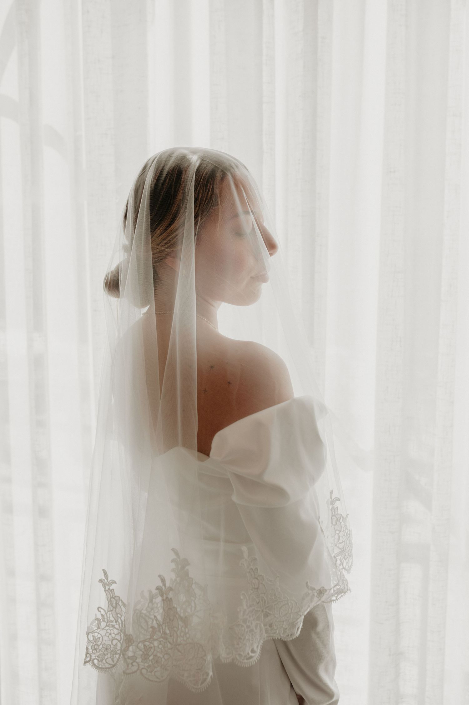 Ethereal bridal portraits through white curtains showing a delicate lace veil and wedding gown silhouette.