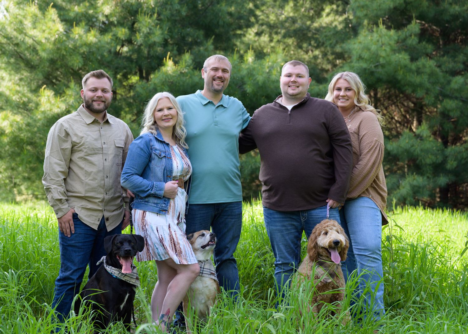 Family group portrait taken outdoors in a lush green field with pine trees.