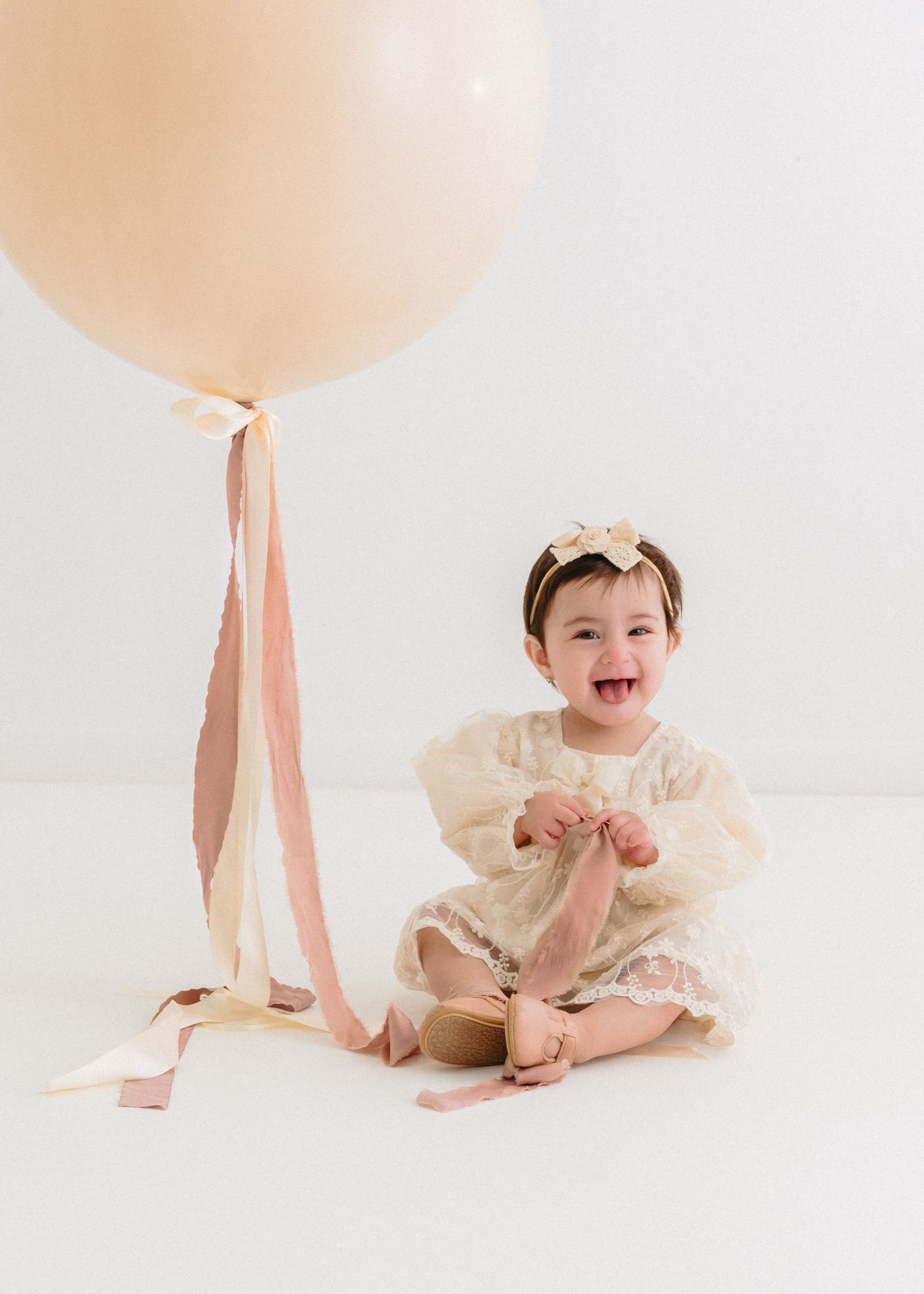 Baby in cream dress playing with large peach balloon with ribbons. Appleton, Green Bay & Kaukauna