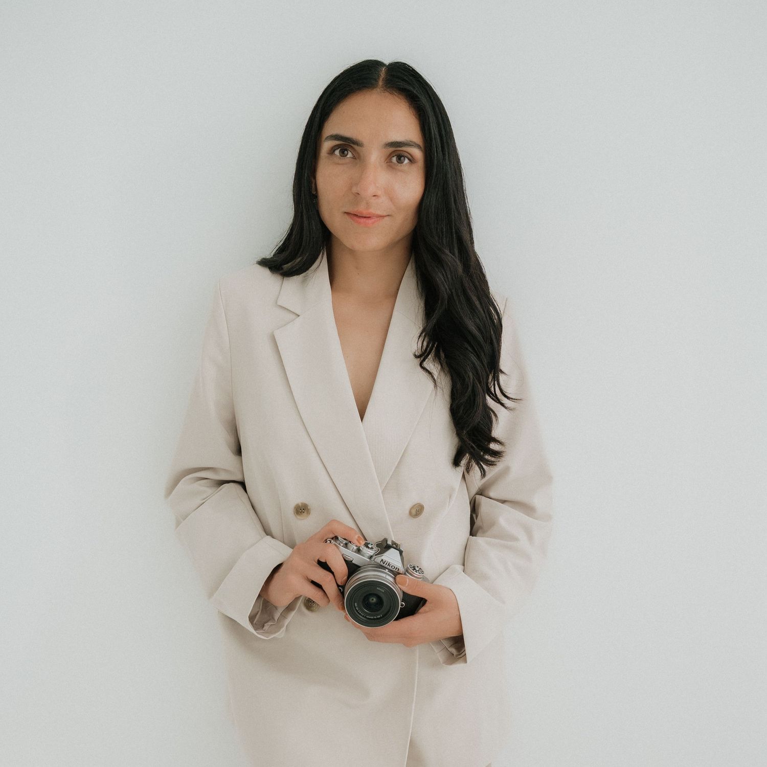 Woman in light colored suit holding camera against minimalist white background. Appleton, Green Bay & Kaukauna