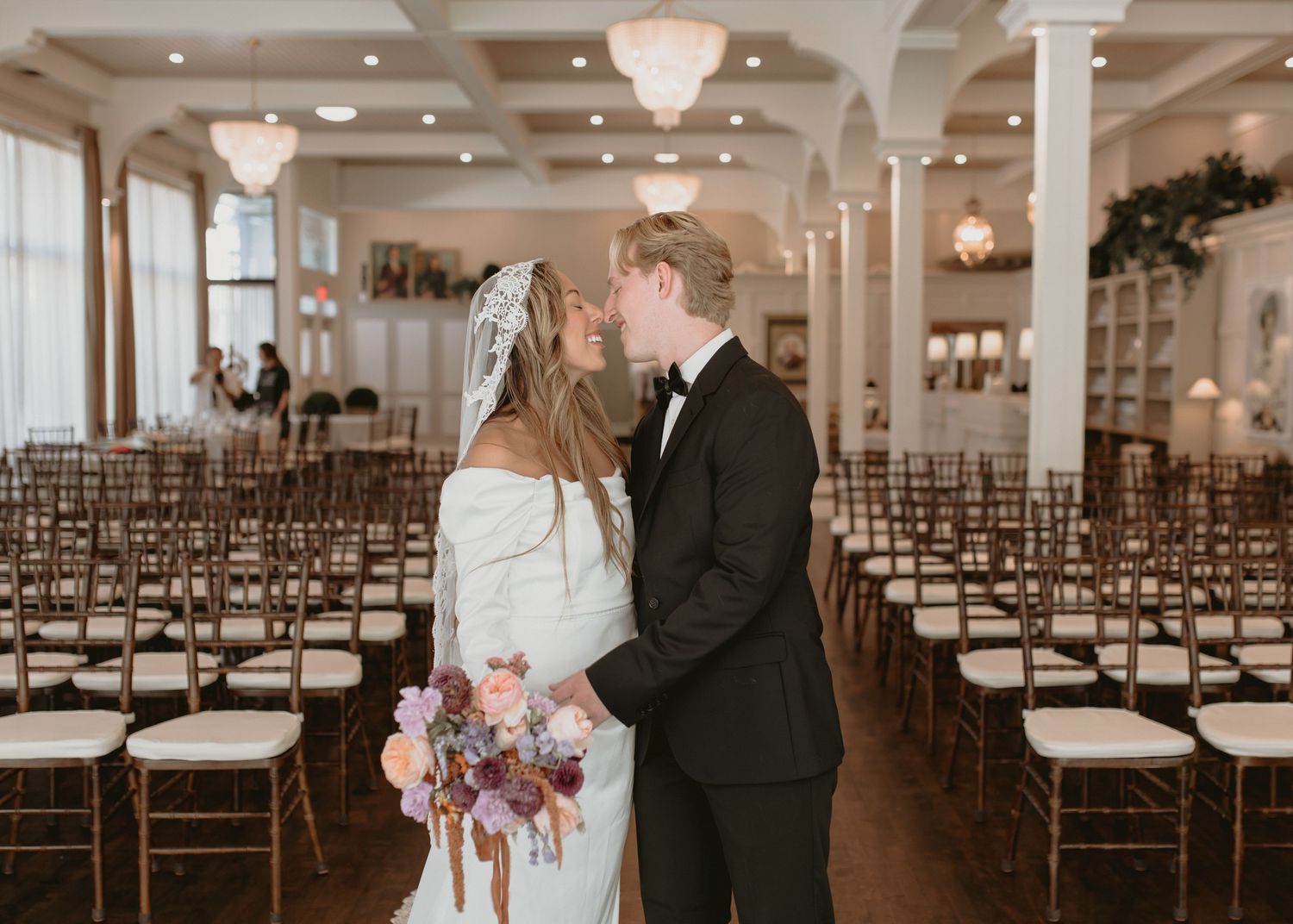 A couple shares a romantic moment in a grand ballroom with crystal chandeliers and rows of chairs.