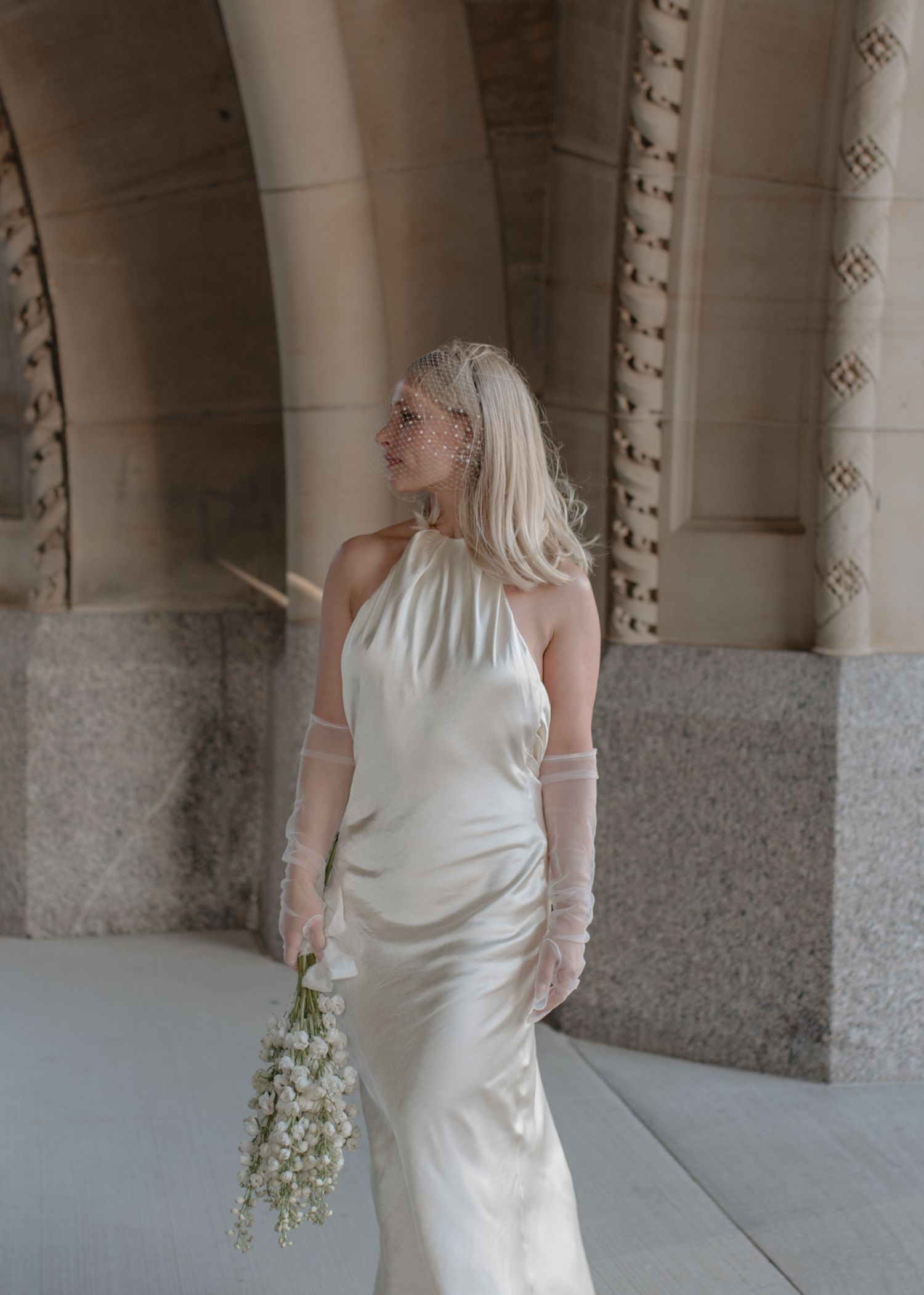 Elegant bride in white halter dress holding white floral bouquet poses in front of stone archway columns.