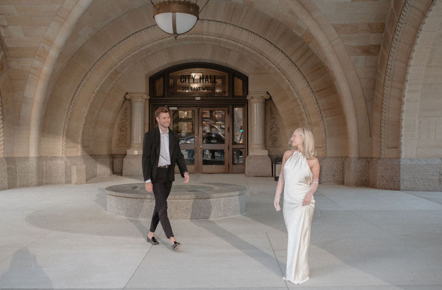 Couple in formal attire walks beneath grand arched entrance of historic stone building.