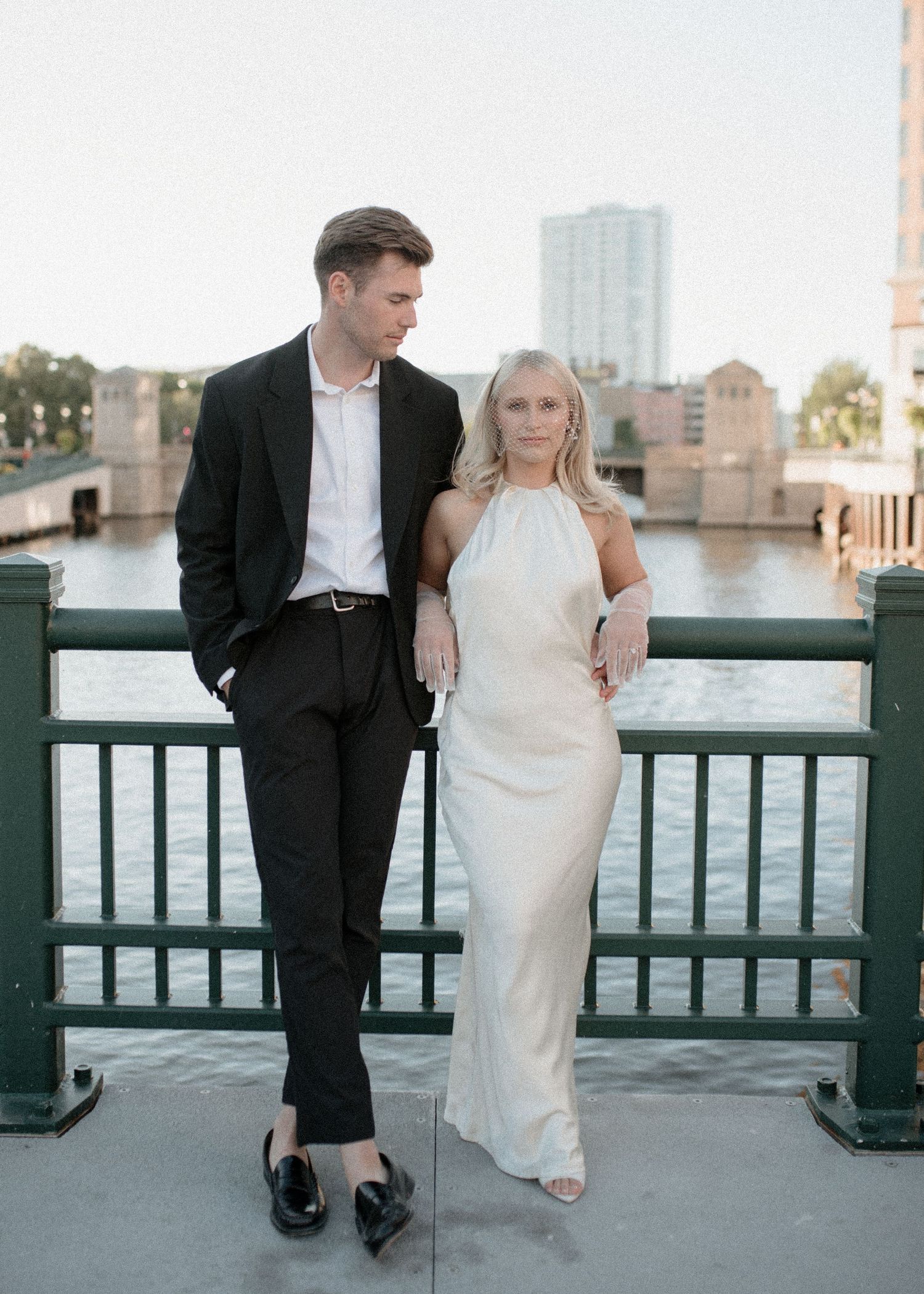 Couple in formal wear poses on bridge railing with cityscape in background.