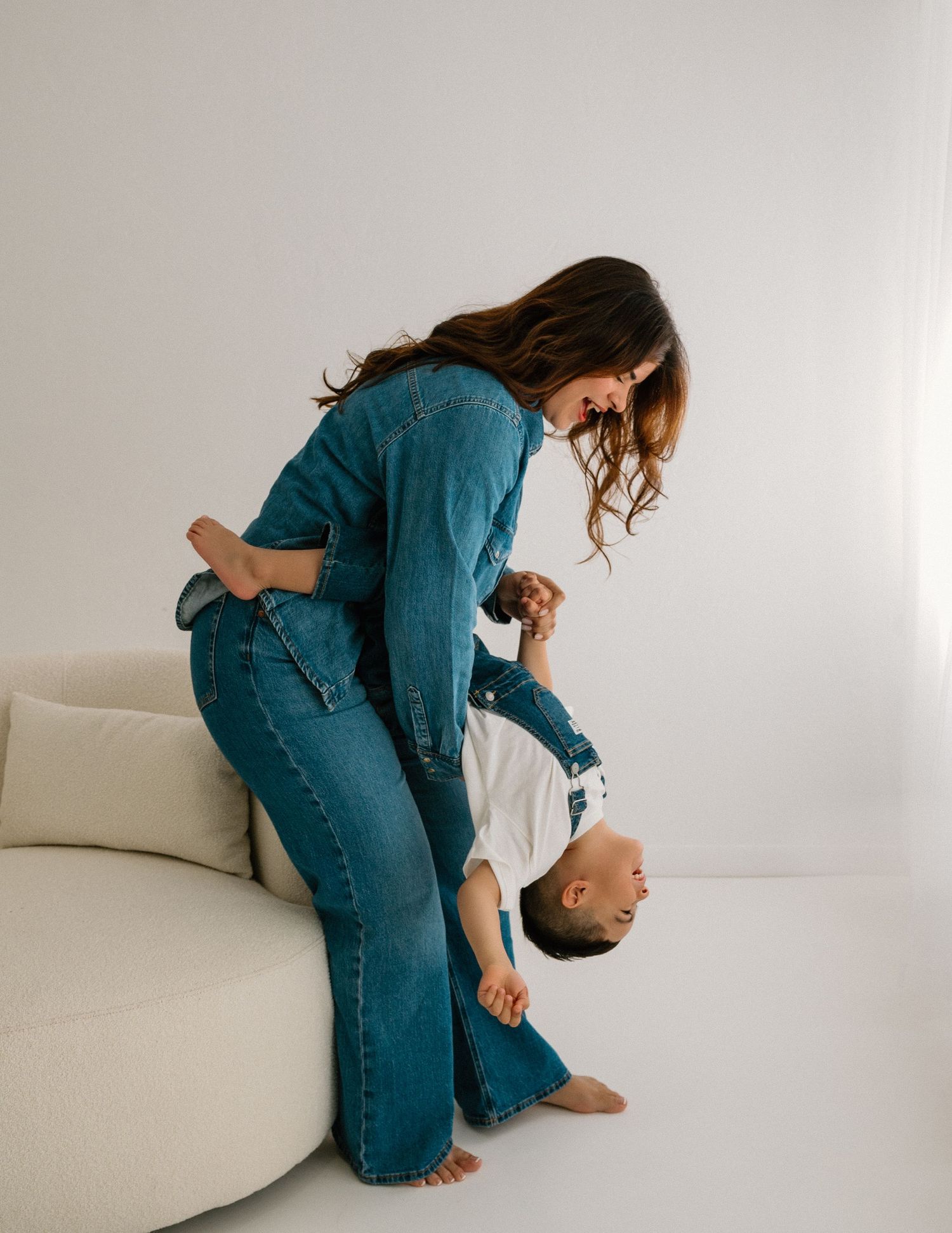 Woman in denim plays and poses with baby during casual indoor lifestyle photoshoot.