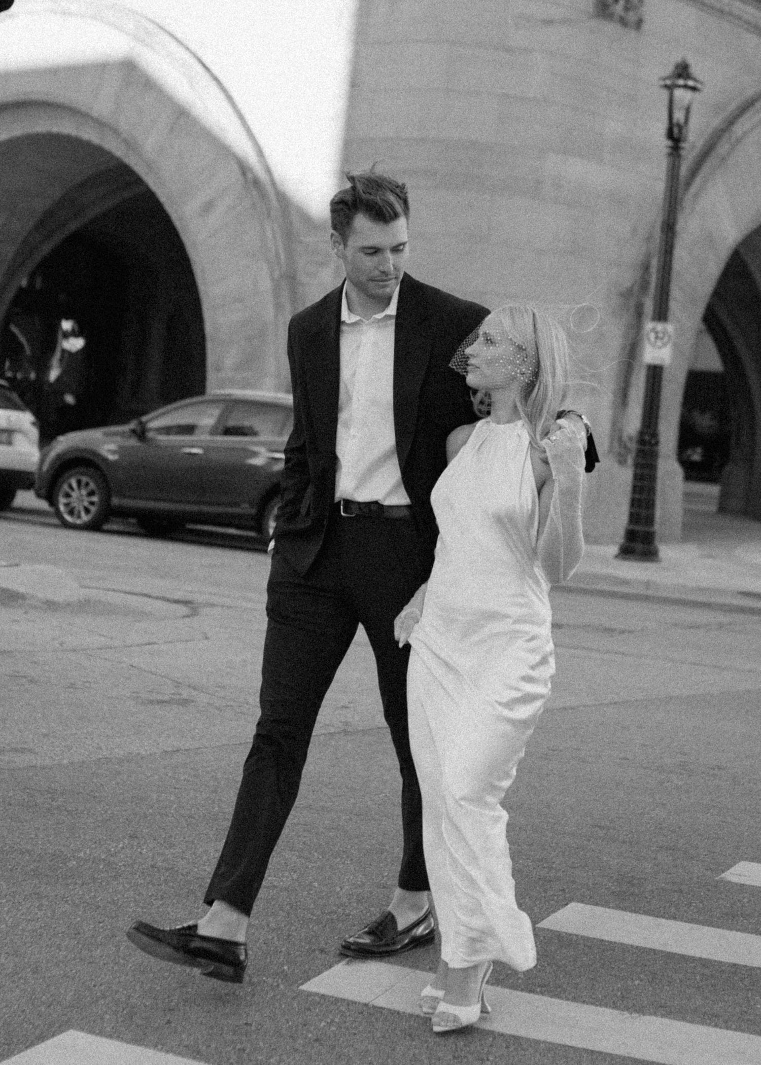 Couple in contrasting formal wear share a laugh together outside historic building with arches.