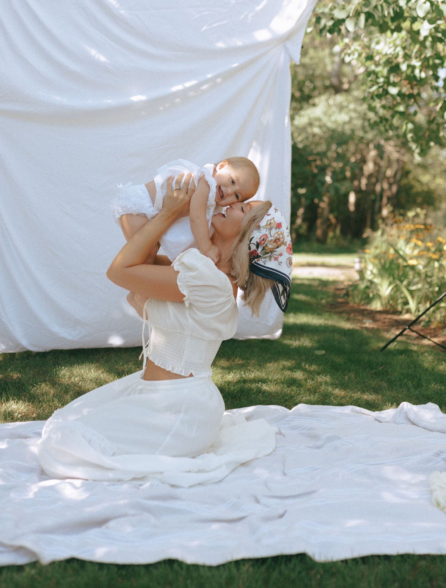 Parent and infant pose together on flowing white fabric during sunlit outdoor session.