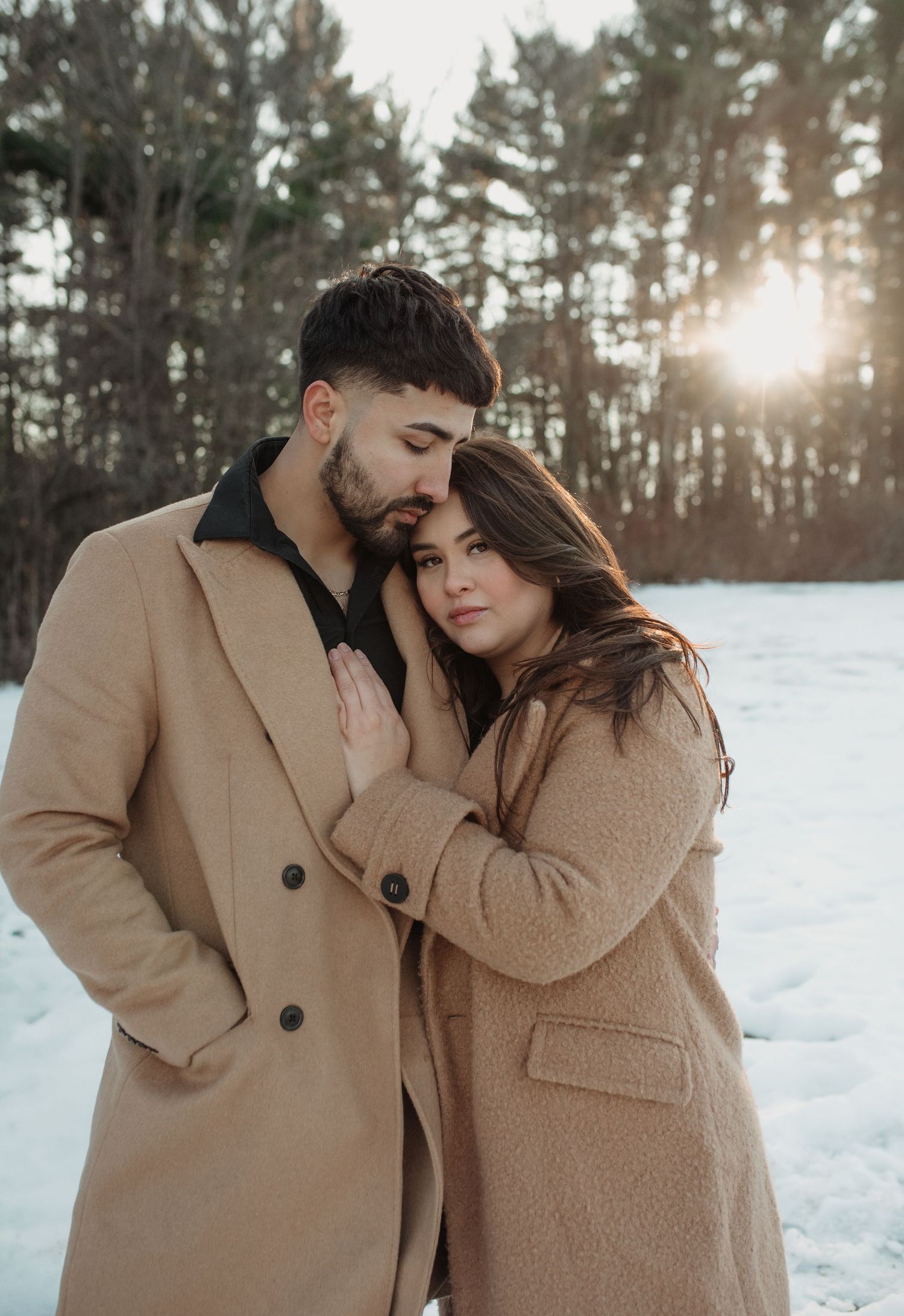 Couple in matching beige coats embracing in a snowy winter forest setting.