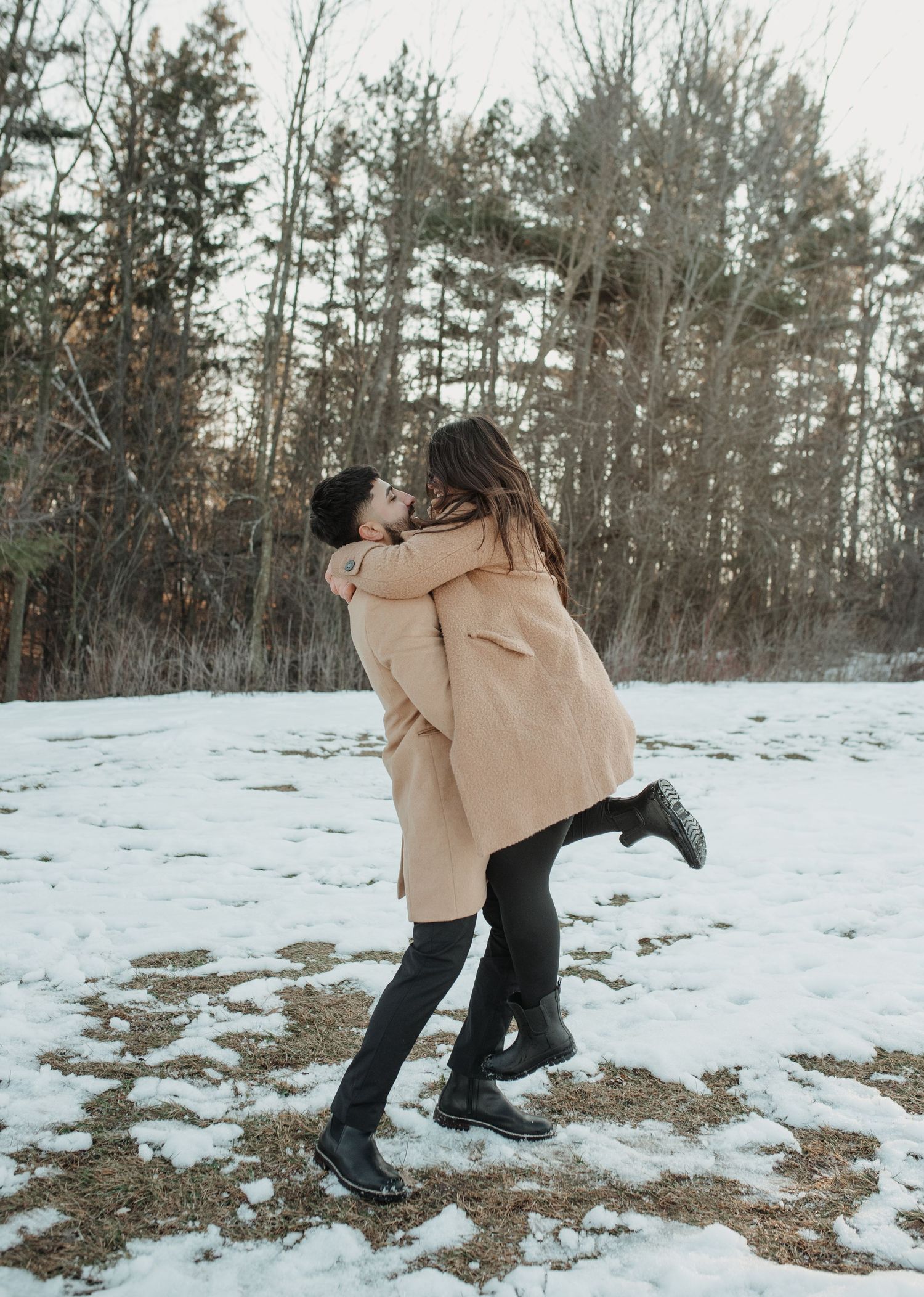 Couple dances playfully in the snow wearing winter coats with forest backdrop. Appleton, Green Bay & Kaukauna