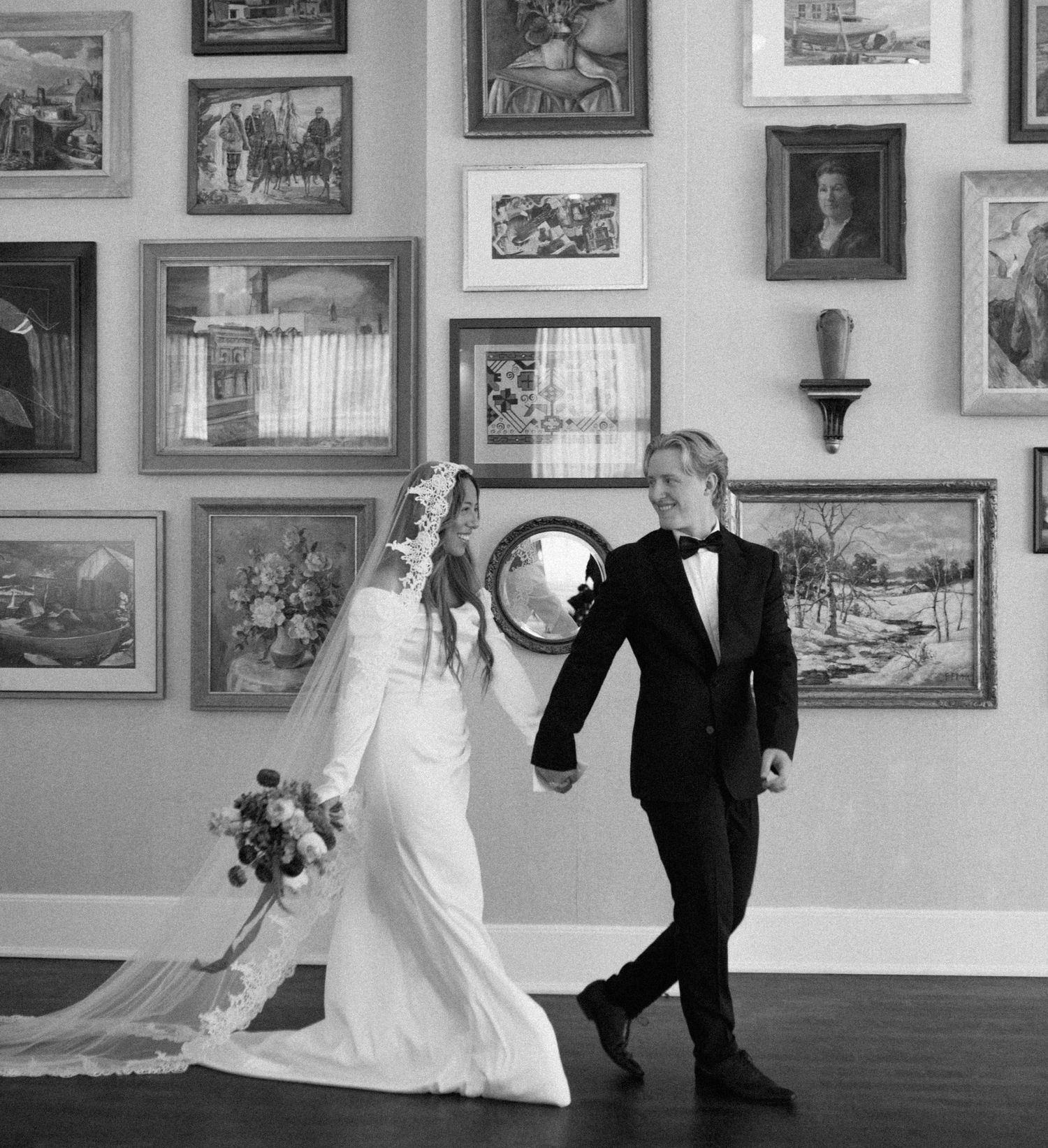 Black and white photo of newlyweds walking through an art gallery filled with framed pictures.