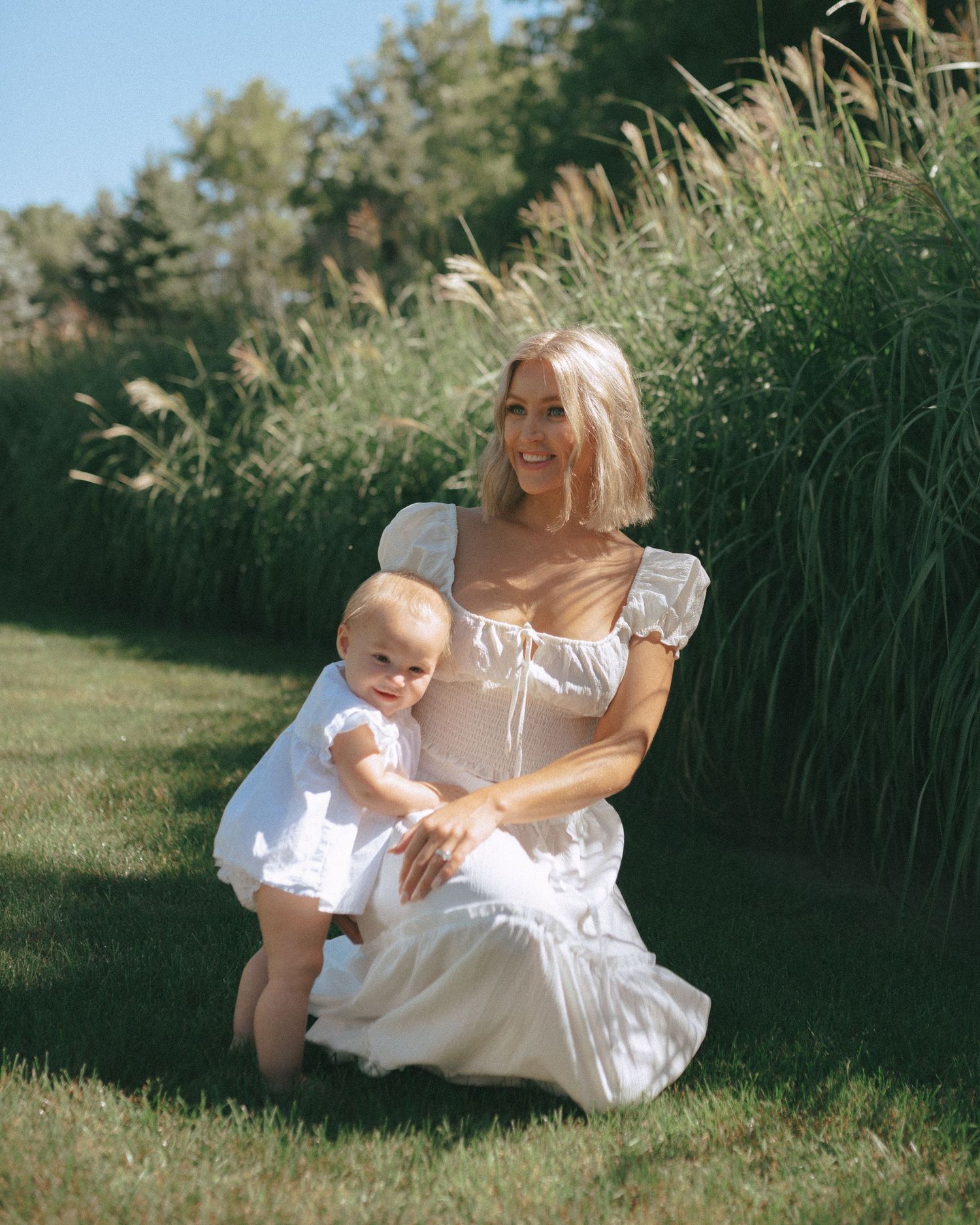 Mother and baby in white dresses pose by tall grasses during summer photoshoot.