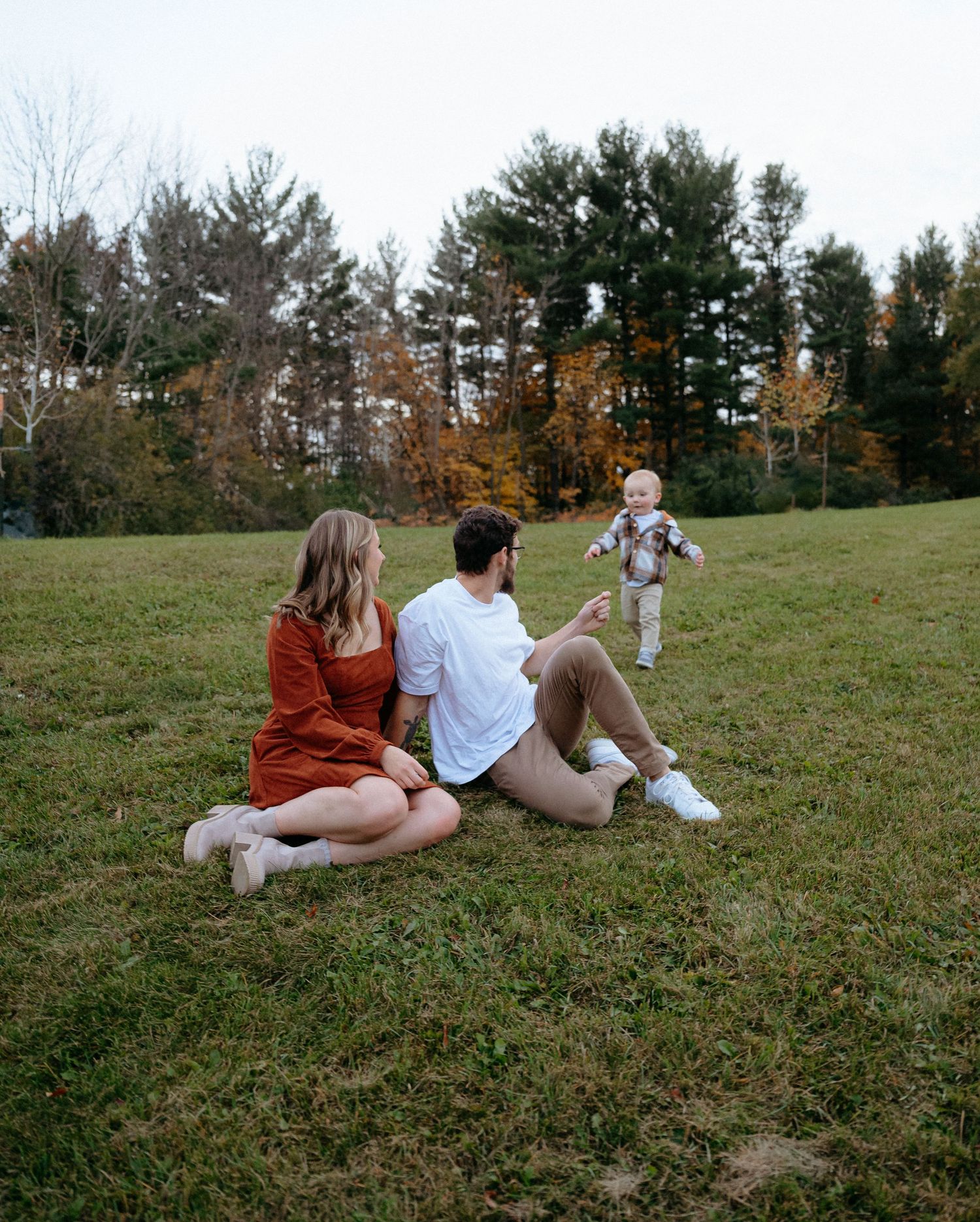 Three figures relax together on a grassy hillside with evergreen trees in the background during autumn.