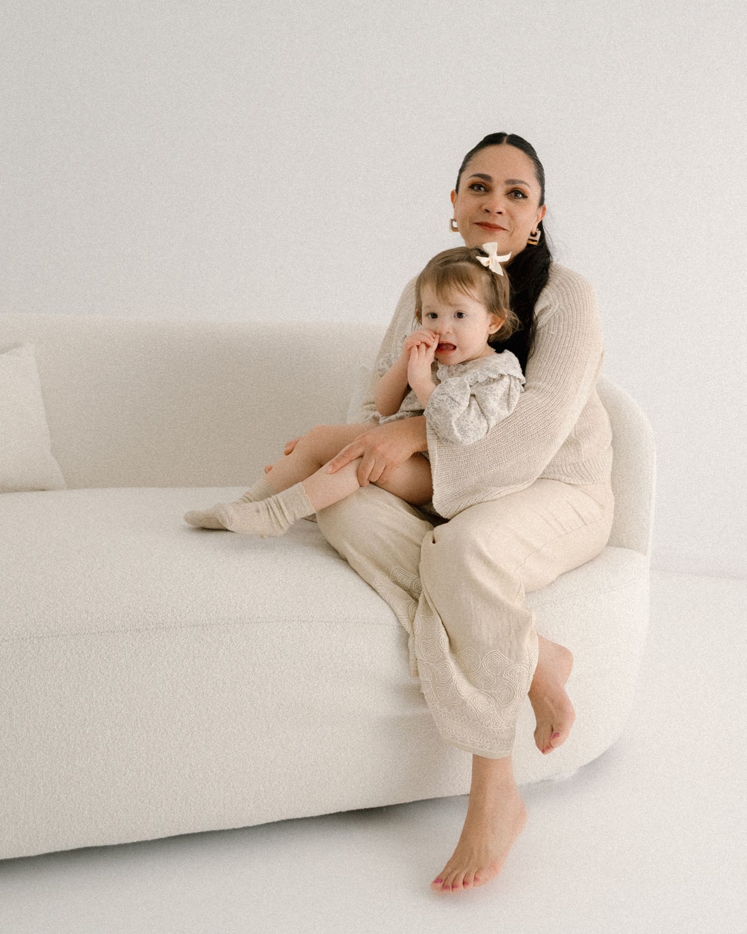 Parent and child in matching cream outfits cuddle on white couch during casual at-home photoshoot.