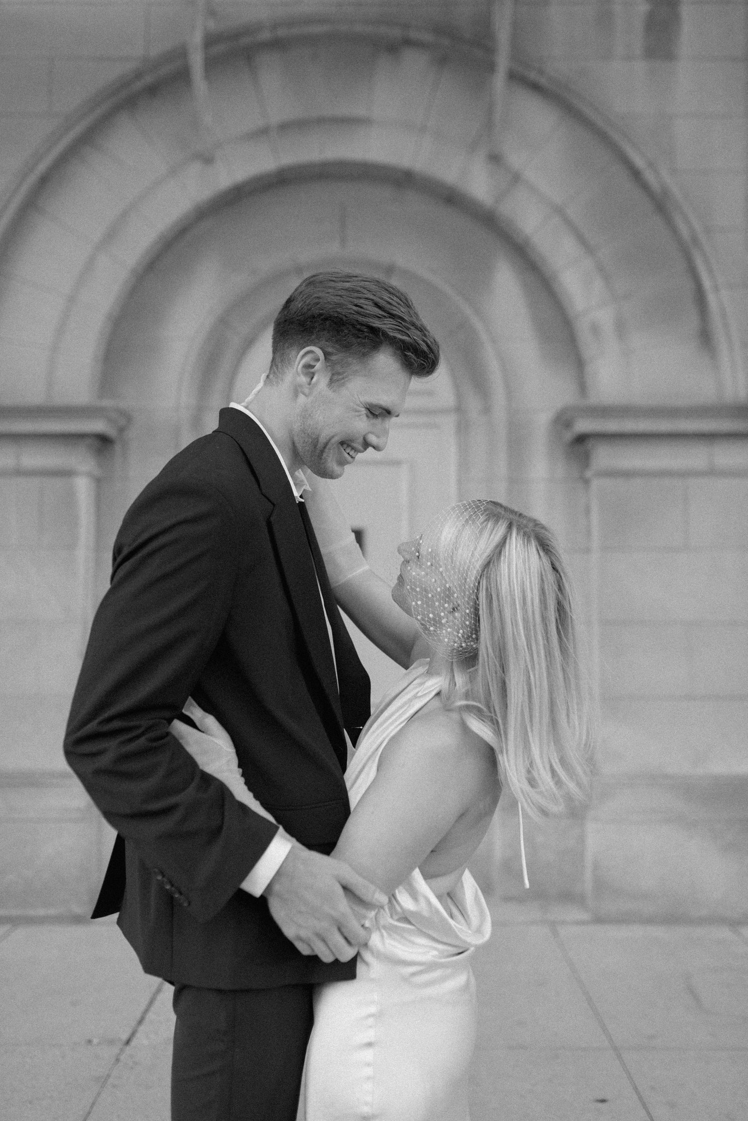 Black and white romantic portrait of couple embracing against architectural backdrop.