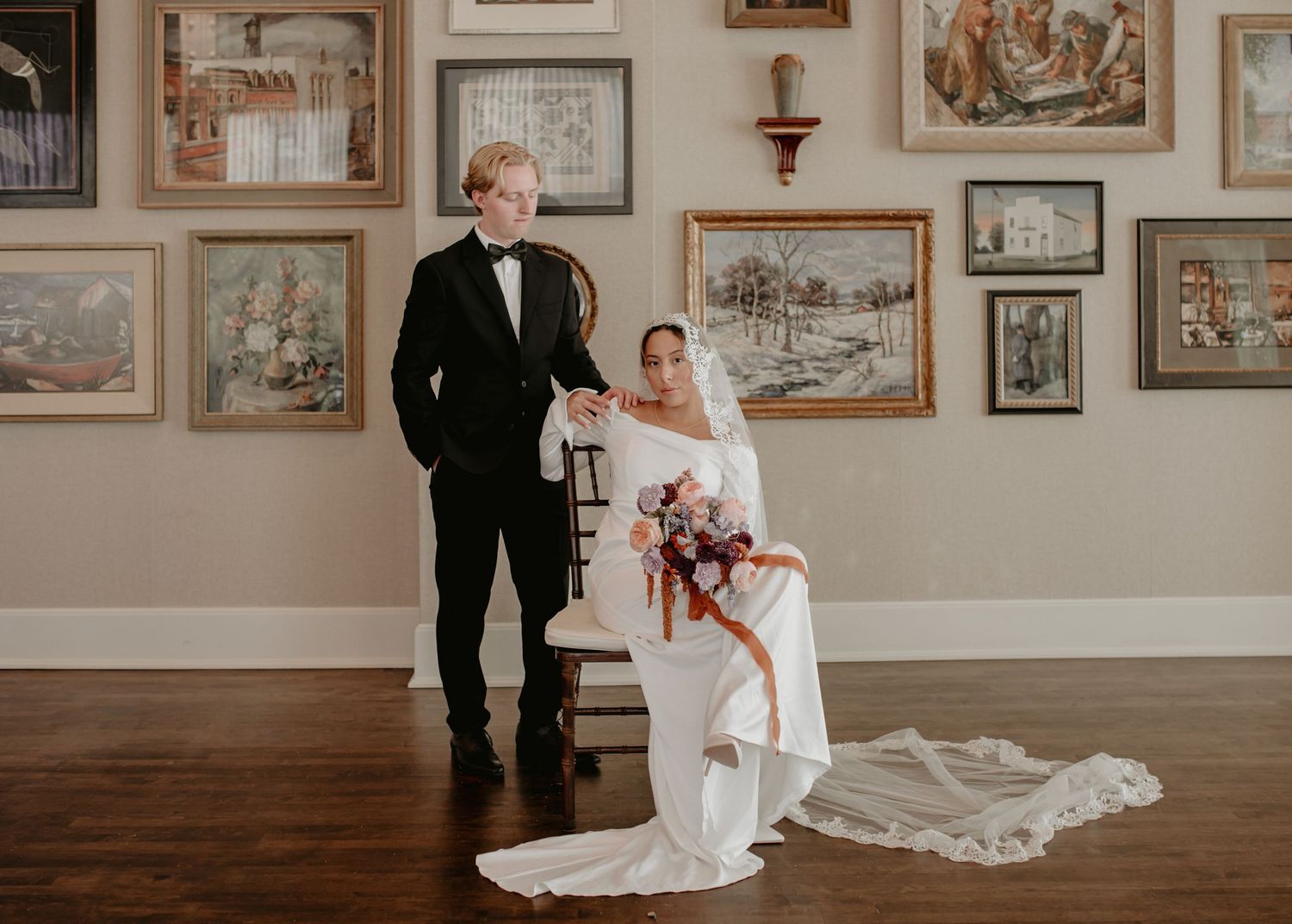 A couple poses in an elegant gallery space surrounded by vintage artwork and warm lighting.